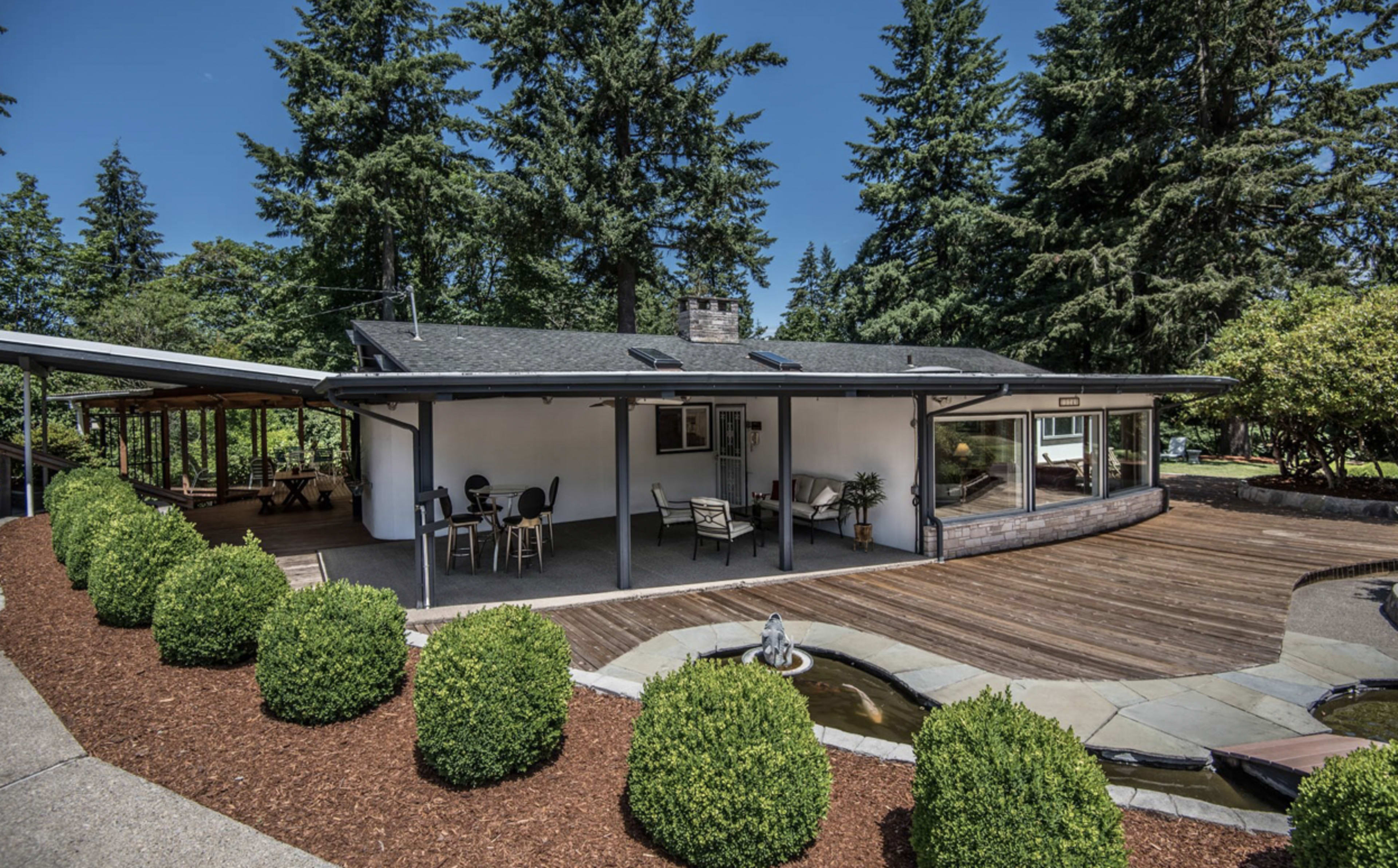 A modern house with a wooden deck and outdoor seating area is surrounded by neatly trimmed shrubs and tall trees.