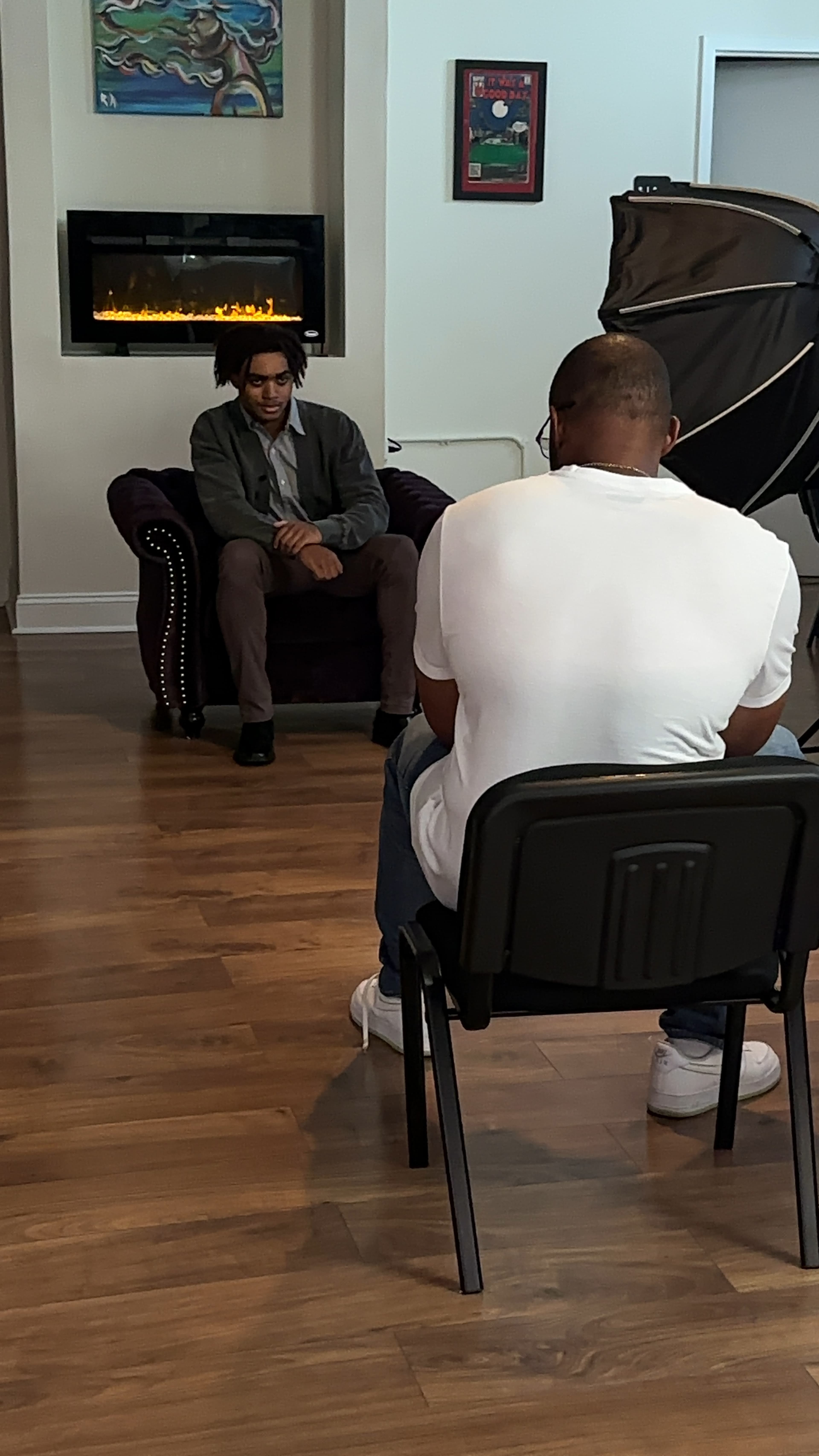 A man sits on a purple armchair while another man takes photographs from a chair in front of him.