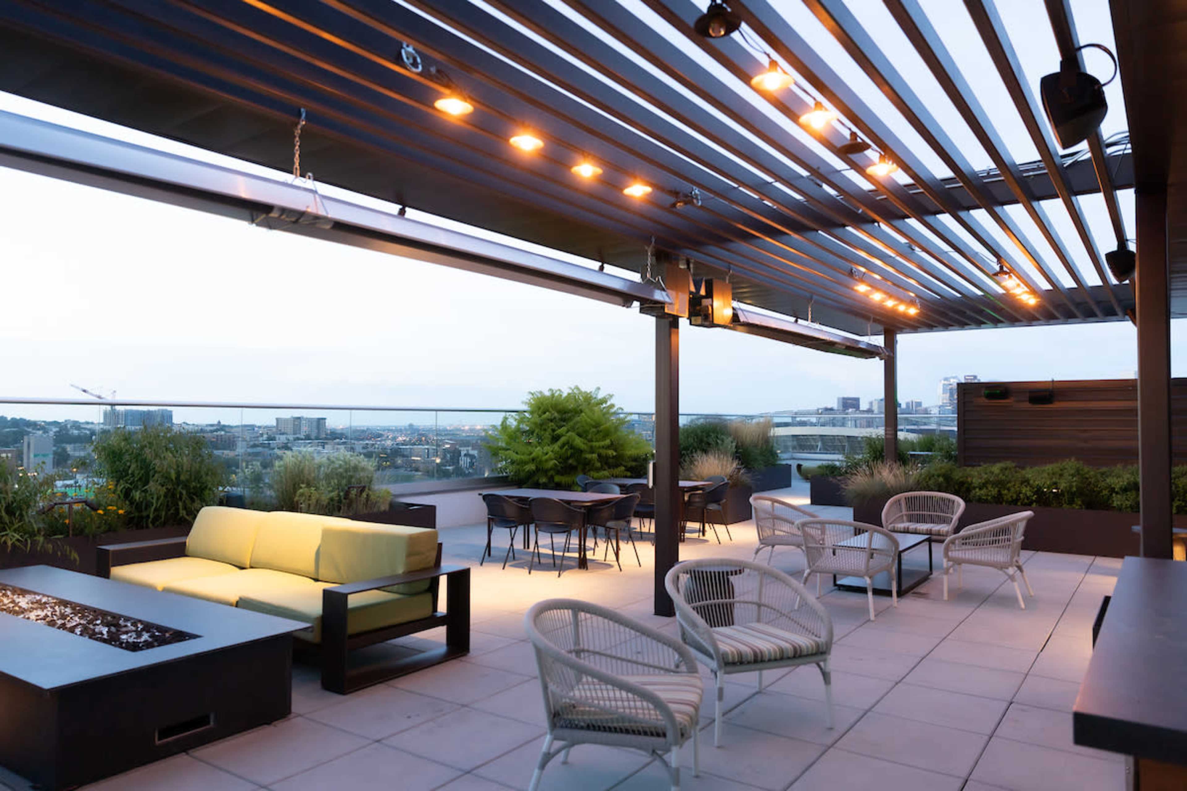 The image shows a rooftop terrace with modern seating, a fire pit, and city views under a striped overhead structure.