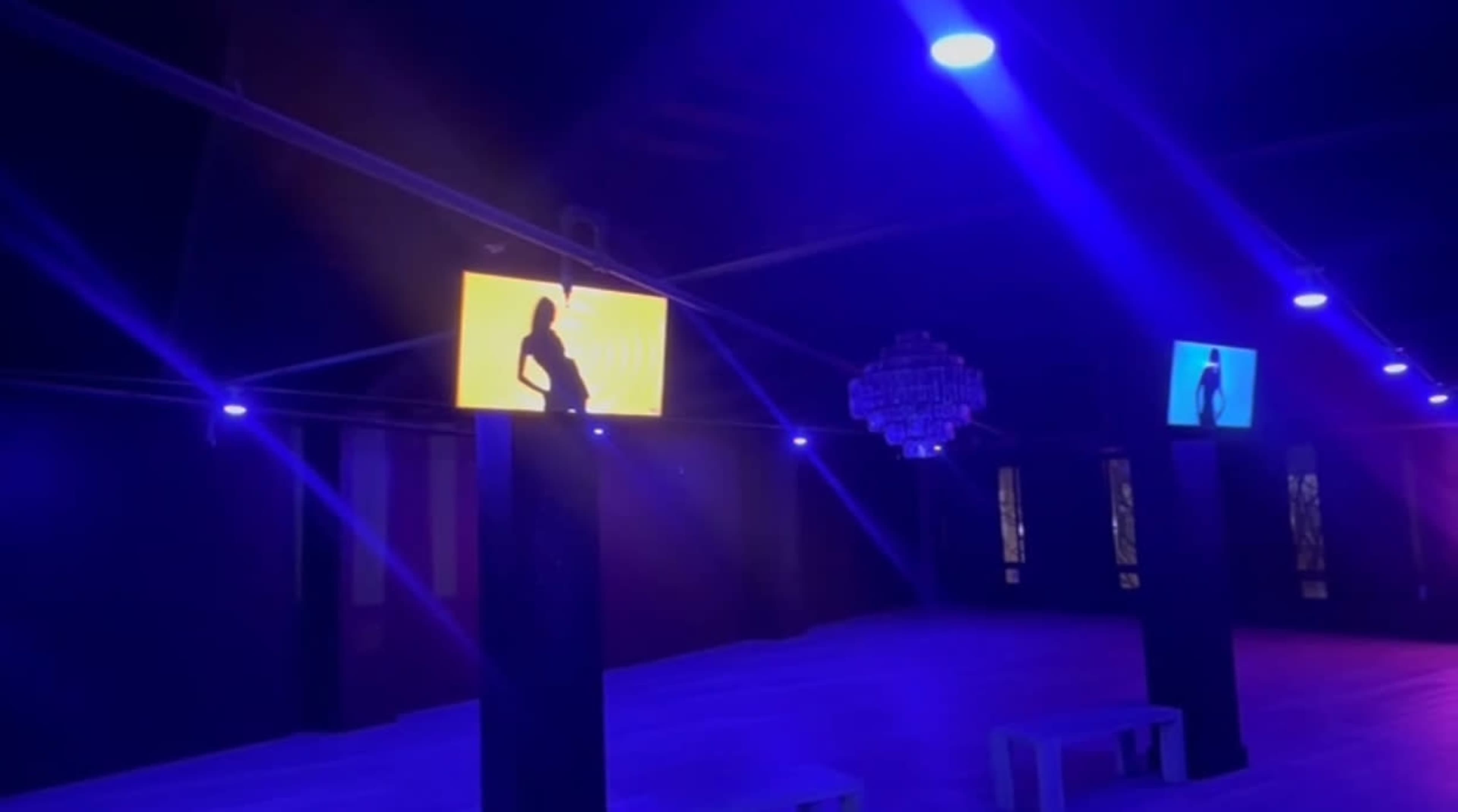 The image shows a dimly lit venue featuring illuminated screens displaying silhouettes against a backdrop of blue lighting.