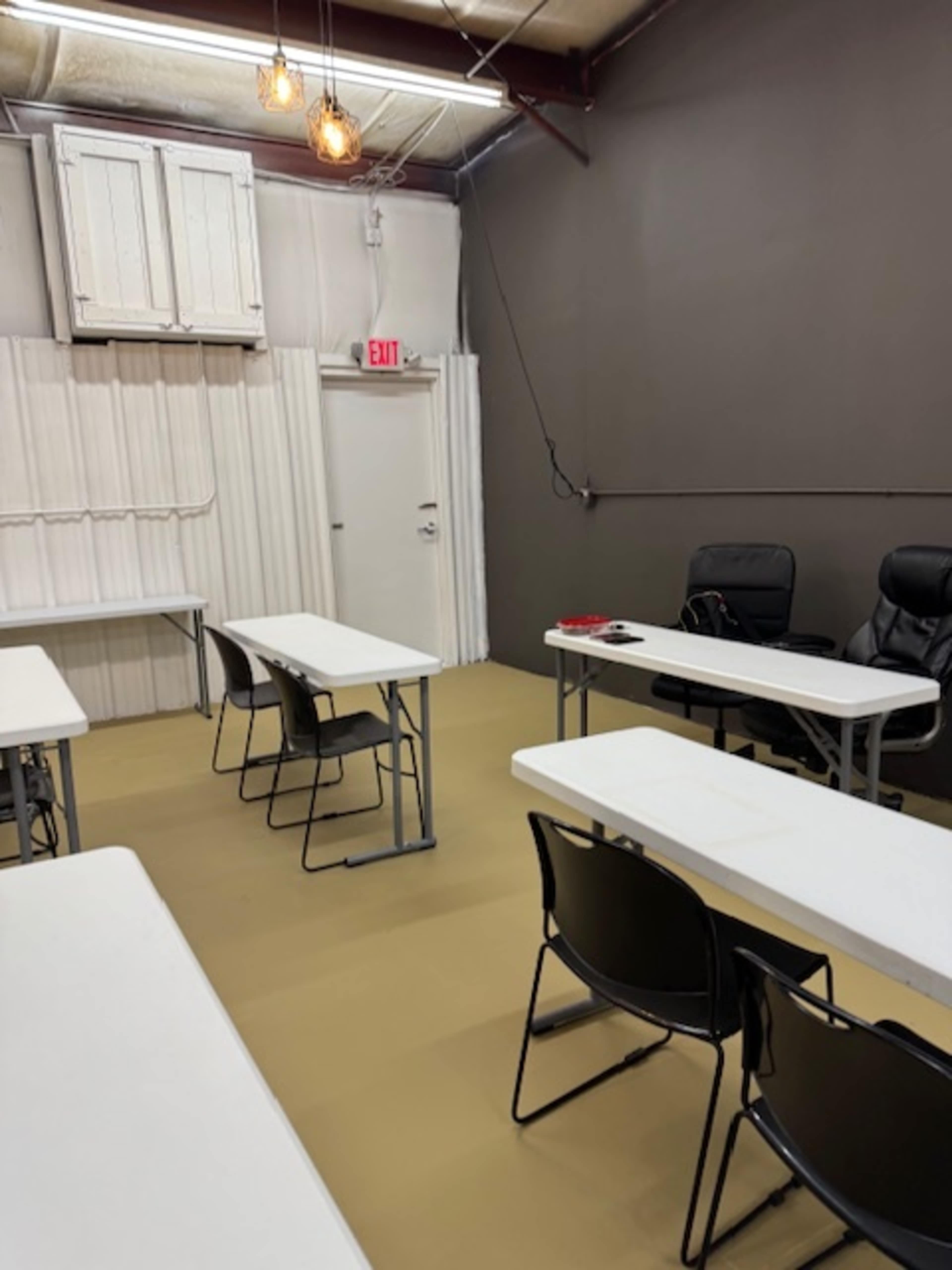 The image shows a sparsely furnished room with several tables and chairs arranged for a meeting, along with an exit door and overhead lighting.