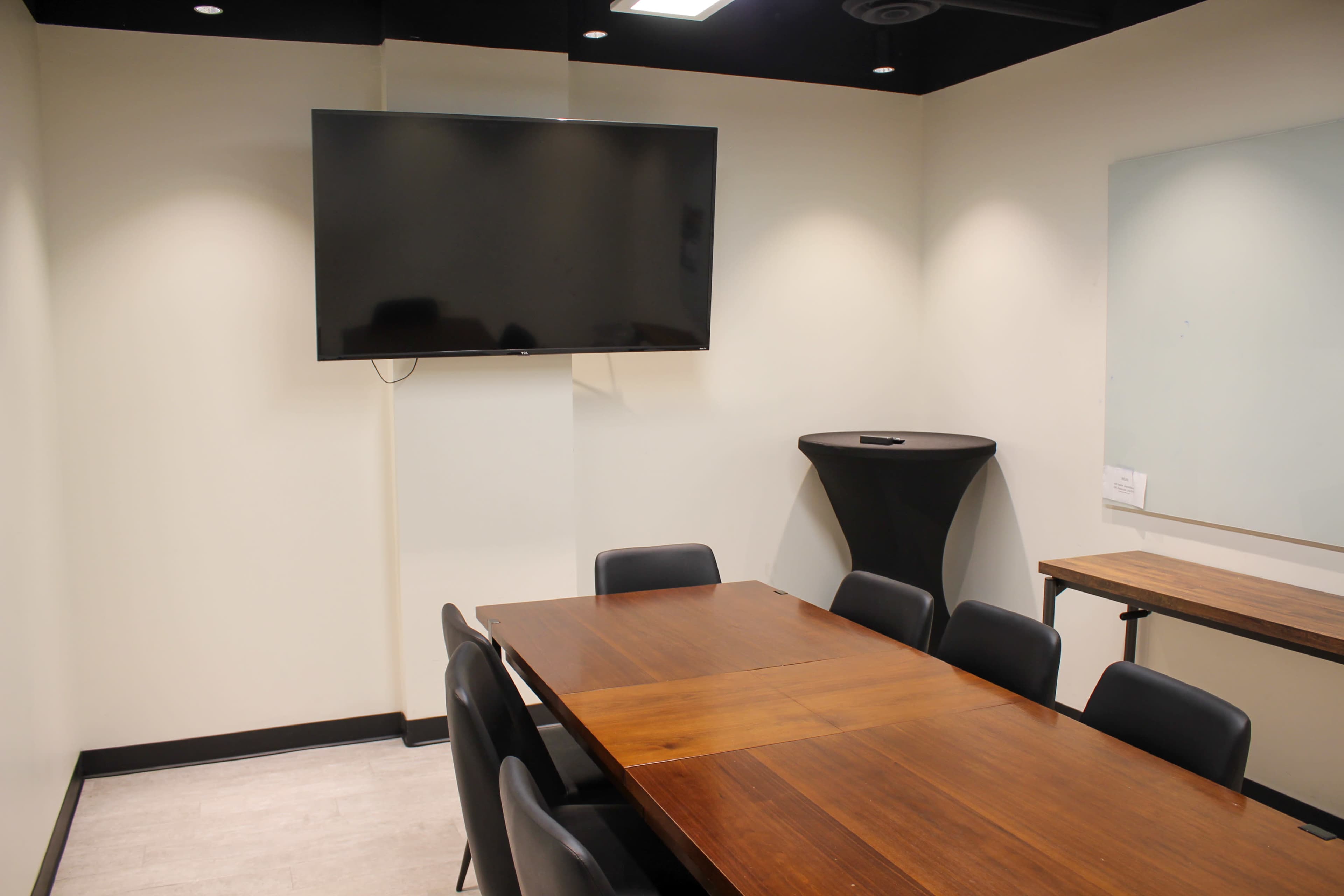 A conference room features a wooden table surrounded by black chairs, with a large flat-screen TV mounted on the wall.