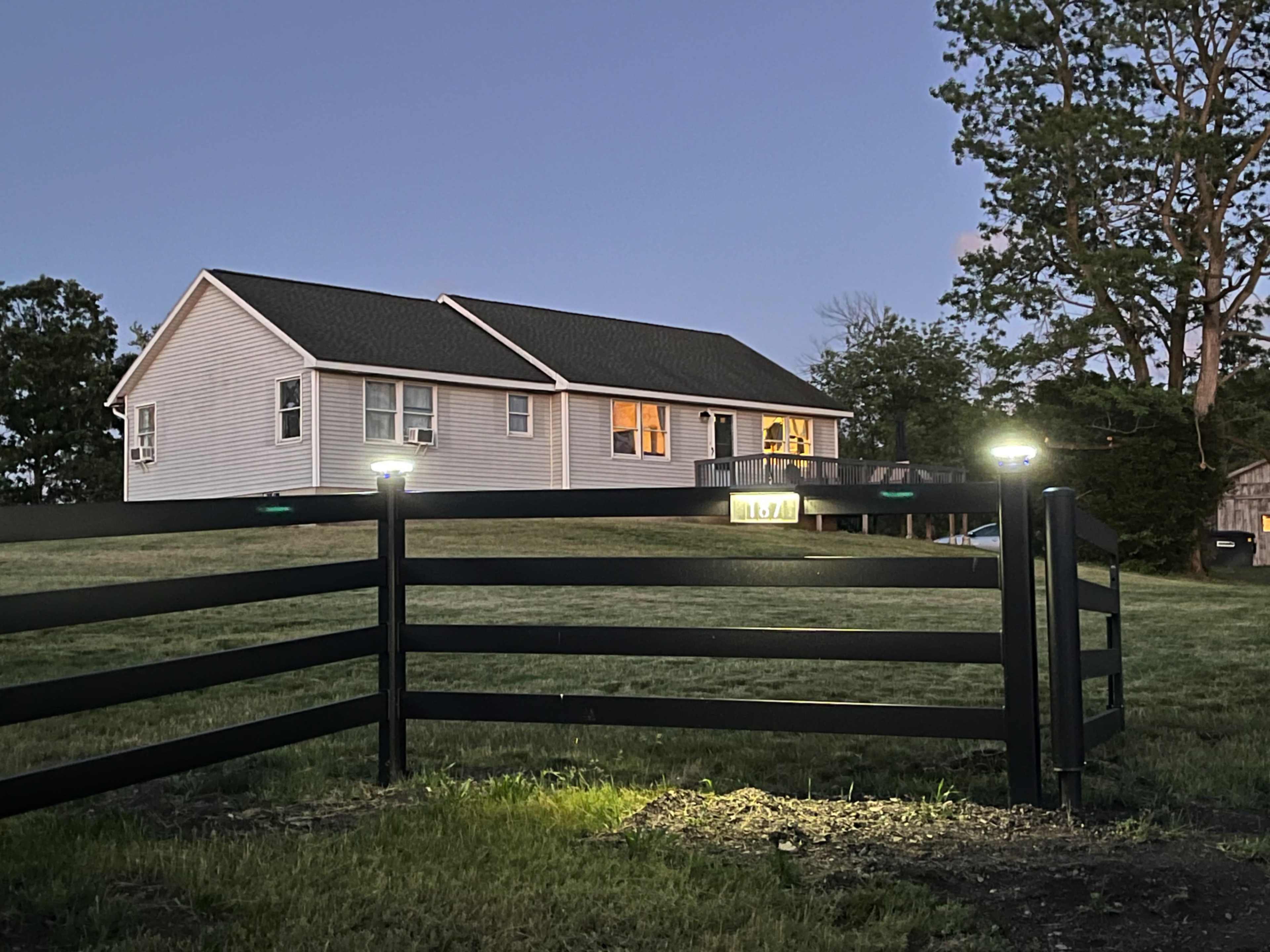 A single-story house with illuminated windows stands behind a black metal fence at dusk.