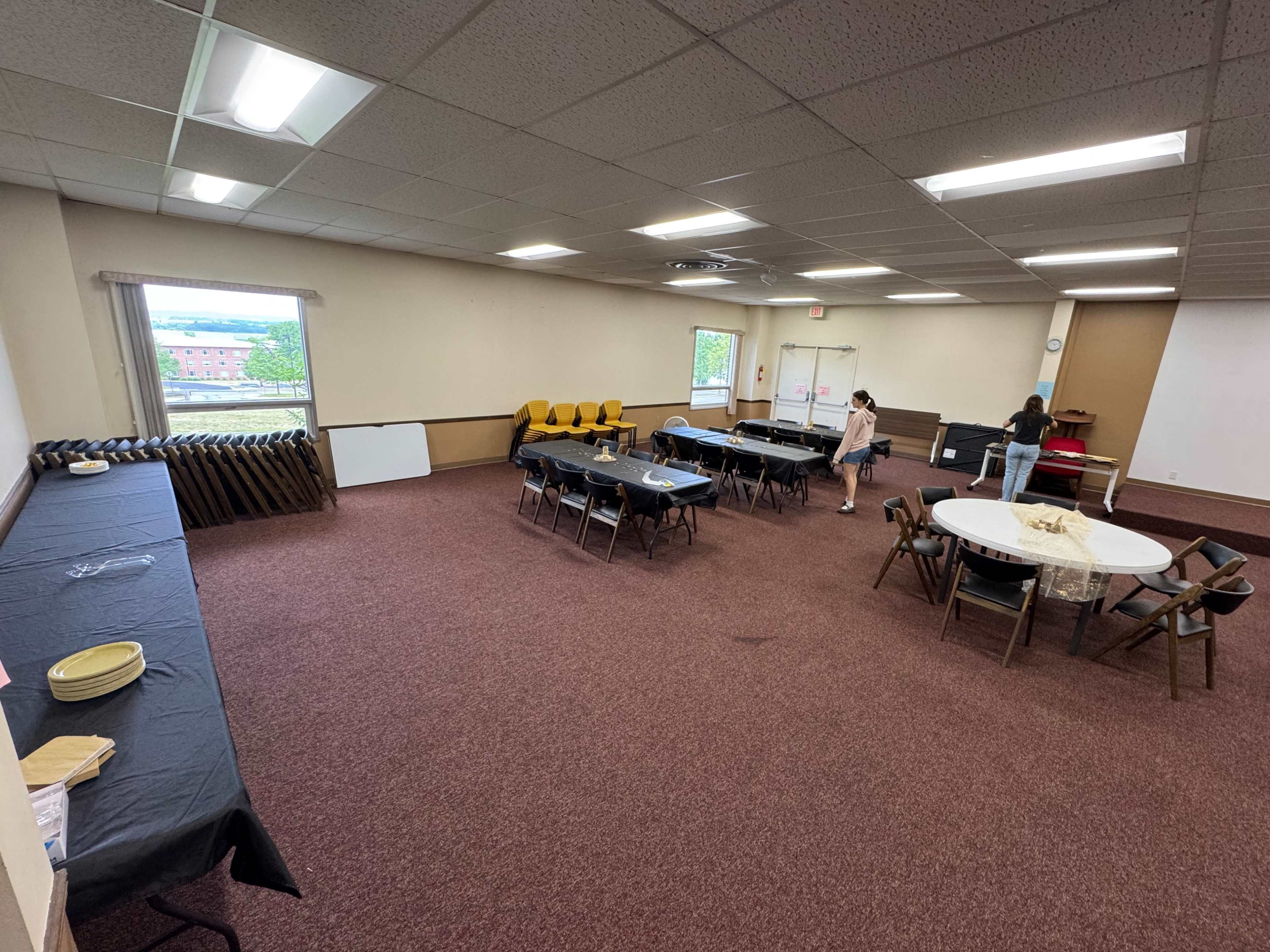 The image shows a large meeting room with several tables arranged in different configurations, a table set against one wall, and a person cleaning in the foreground.