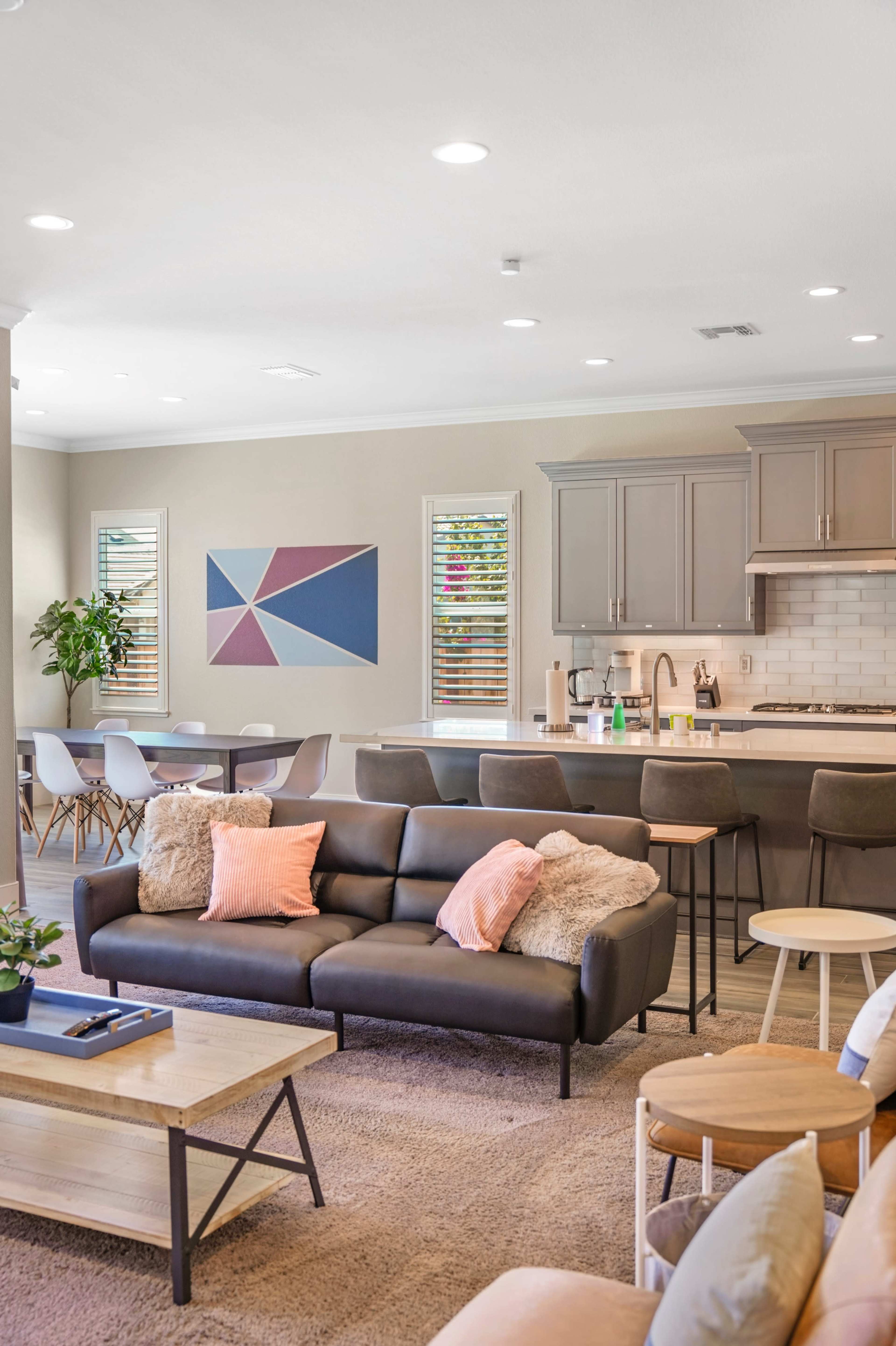 A modern living room features a dark sofa with decorative pillows, a coffee table, and is open to a dining area with a round table and chairs, complemented by a geometric wall art piece.