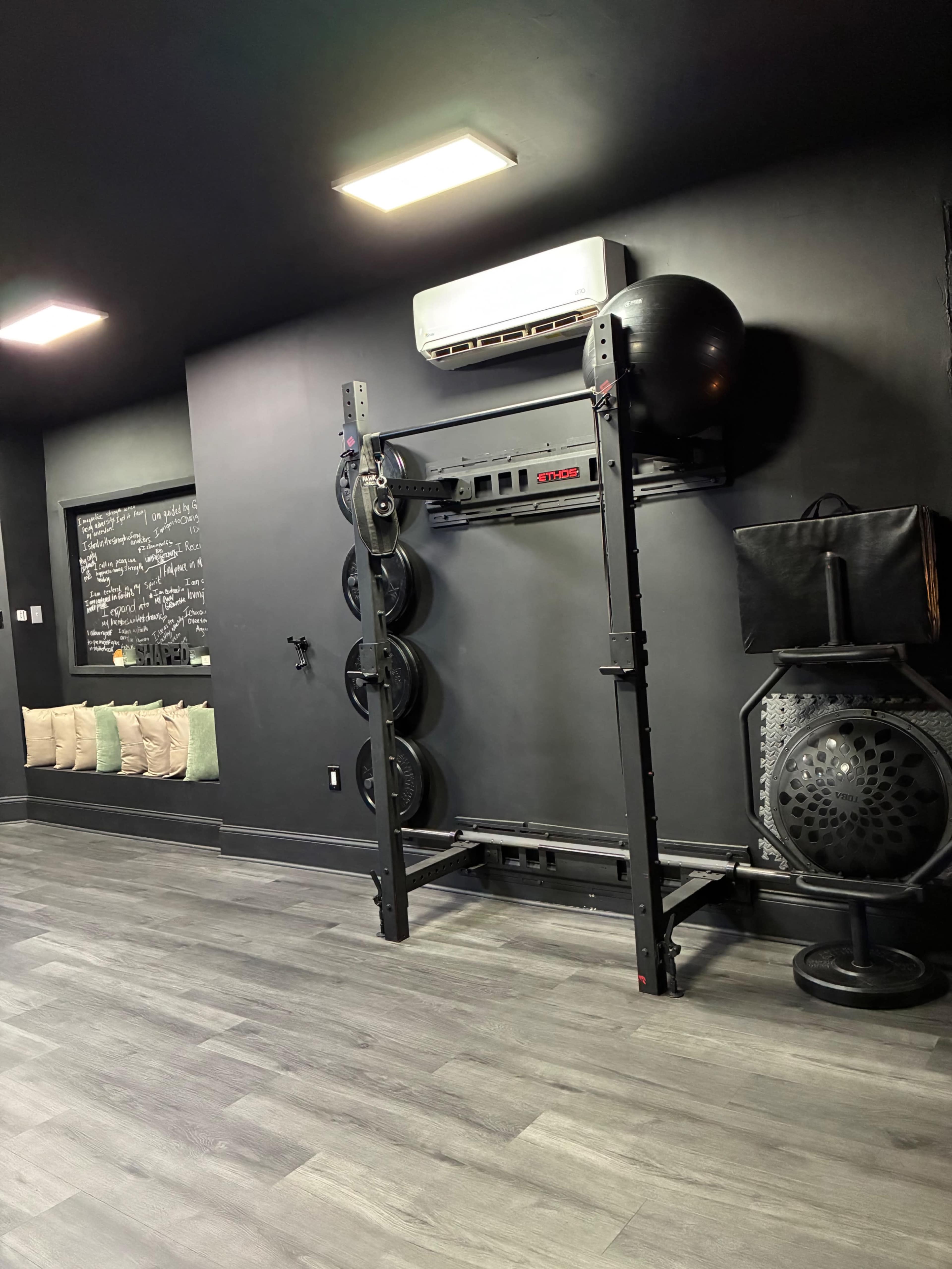 The image shows a dimly lit gym with a black wall, a weight rack equipped with plates, an air conditioning unit, and a seating area with cushions.