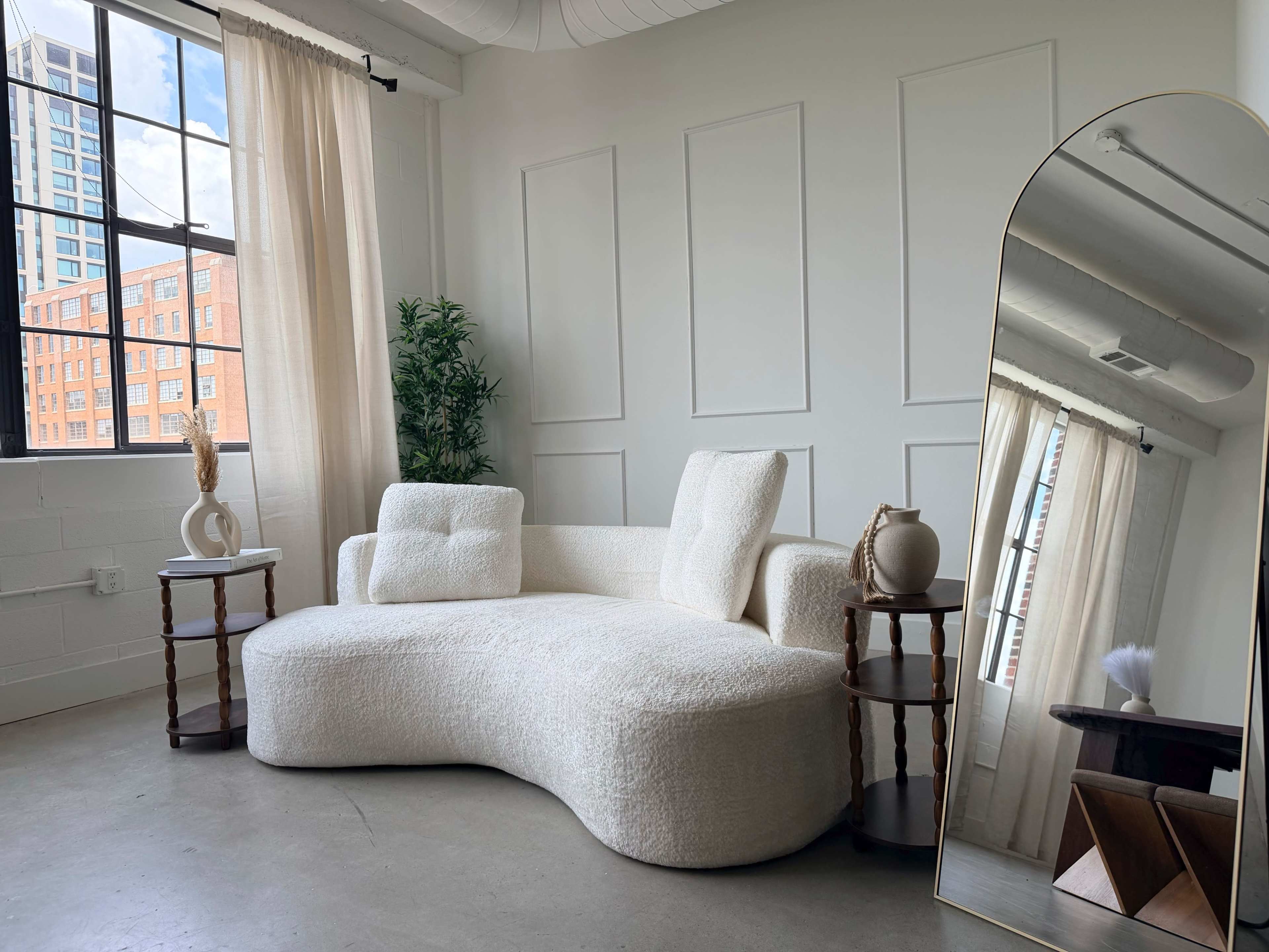 The image shows a modern, minimalist living space featuring a white, curvilinear couch, two small side tables, a large mirror, and a plant near a window with sheer curtains.