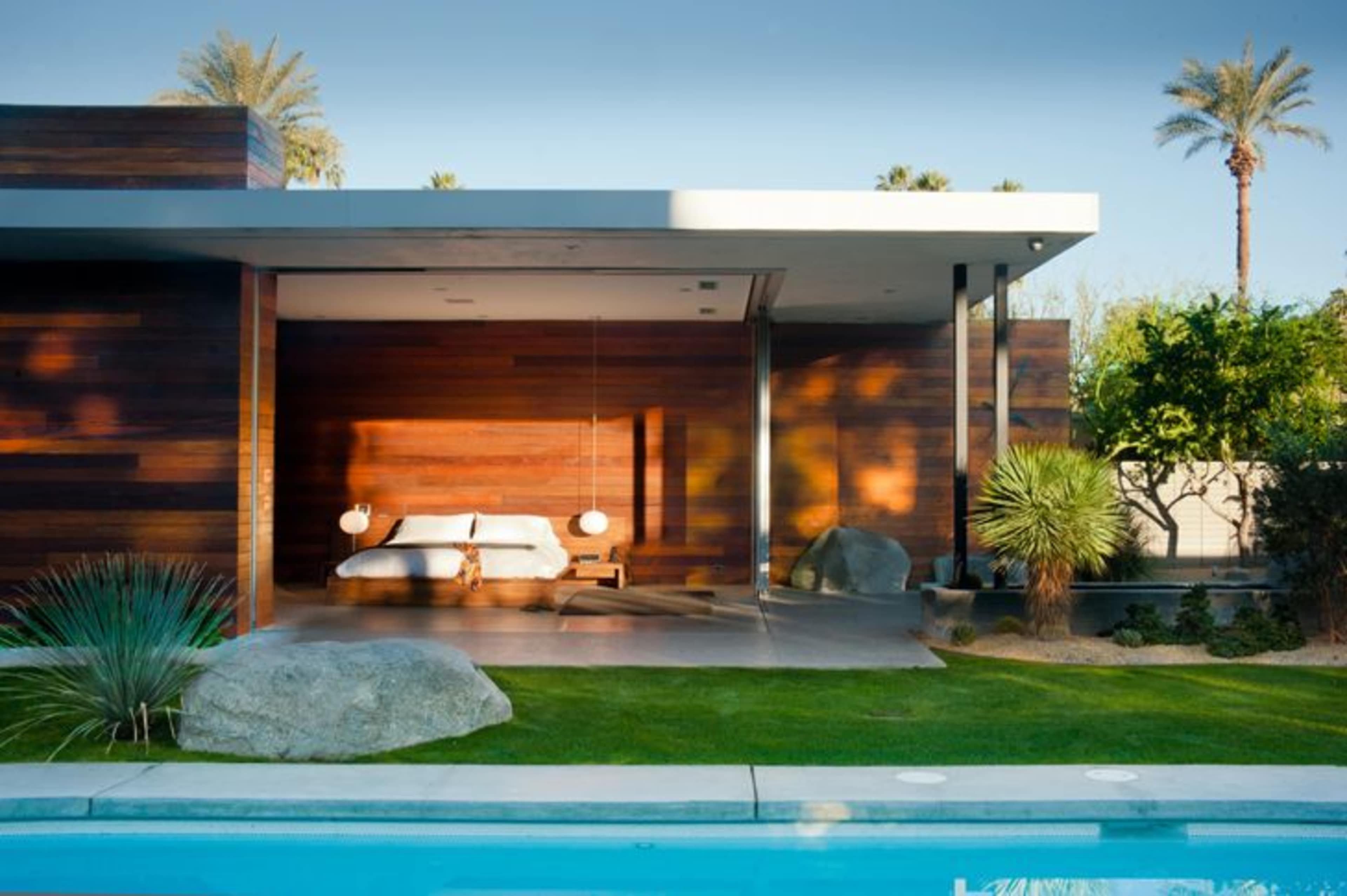 The image shows a modern bedroom with a wooden exterior, featuring a bed, hanging bedside lamps, and a clear view of a pool and landscaped garden.