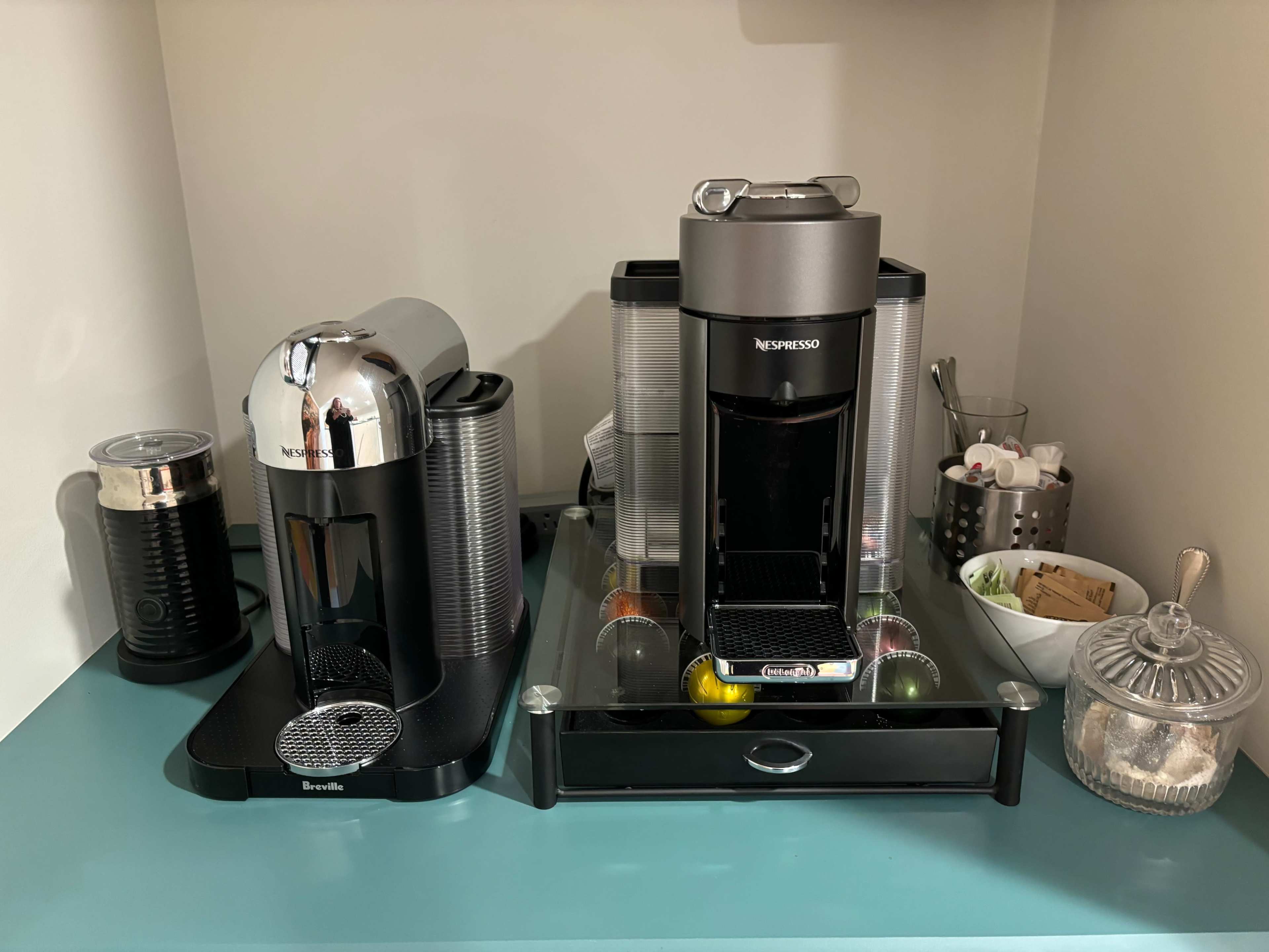 A coffee station featuring a silver Breville espresso machine and a black Nespresso machine, both positioned on a teal countertop alongside various coffee accessories and cups.