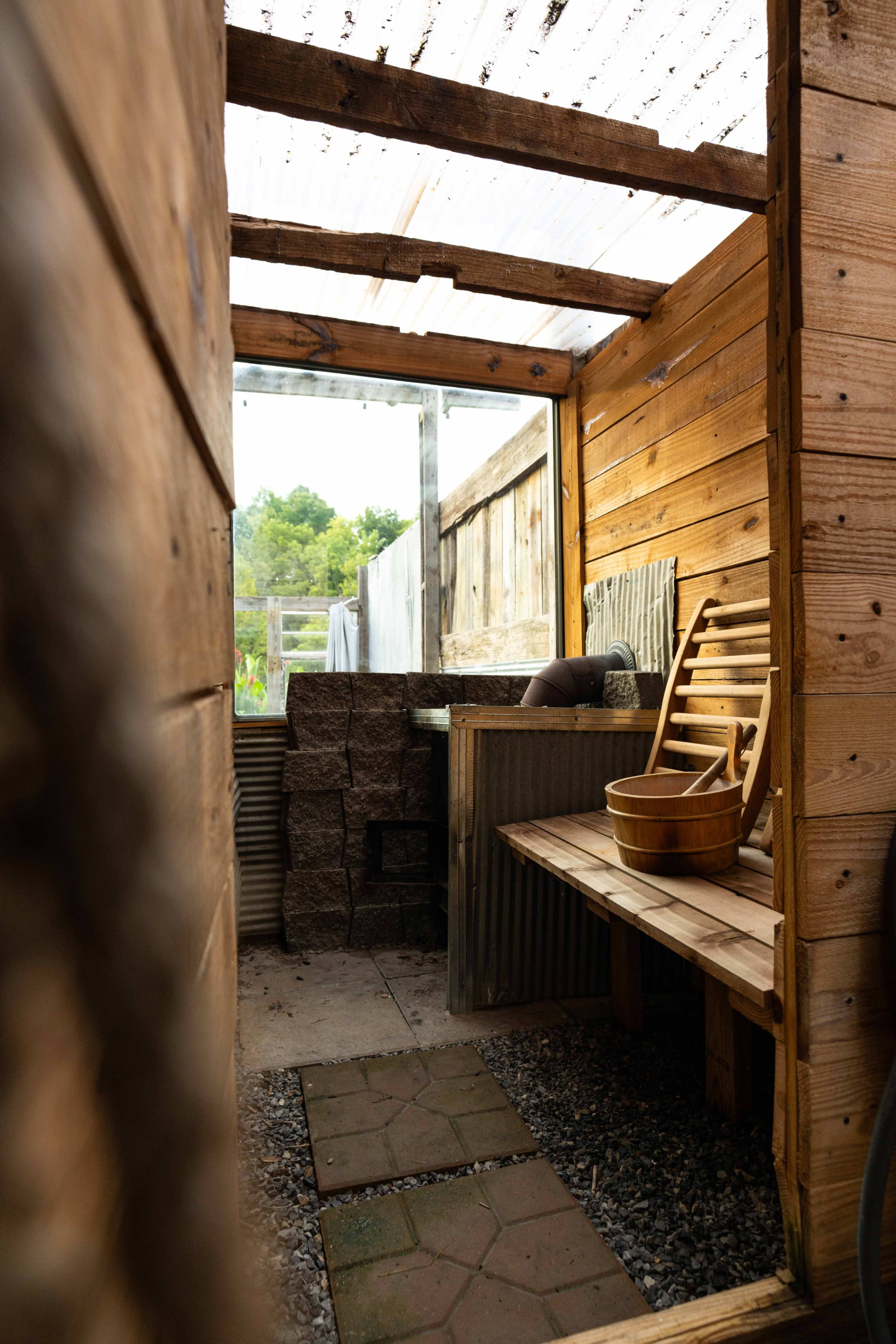 A narrow, wooden structure featuring a bench, stone blocks, and a wooden bucket, with a transparent roof allowing light in.