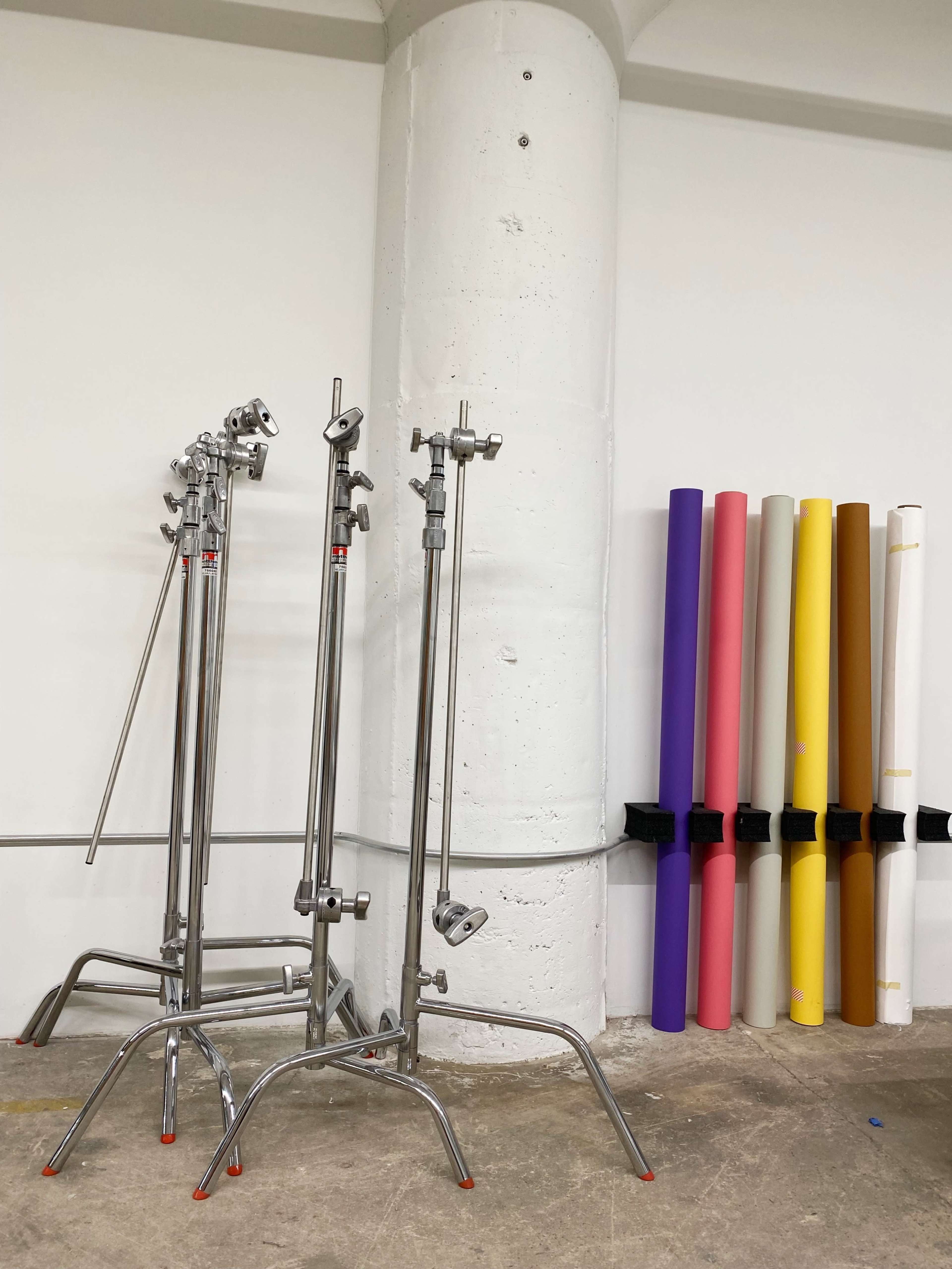 Three silver light stands positioned near a white concrete pillar, with several rolls of backdrop paper in various colors leaned against the wall.