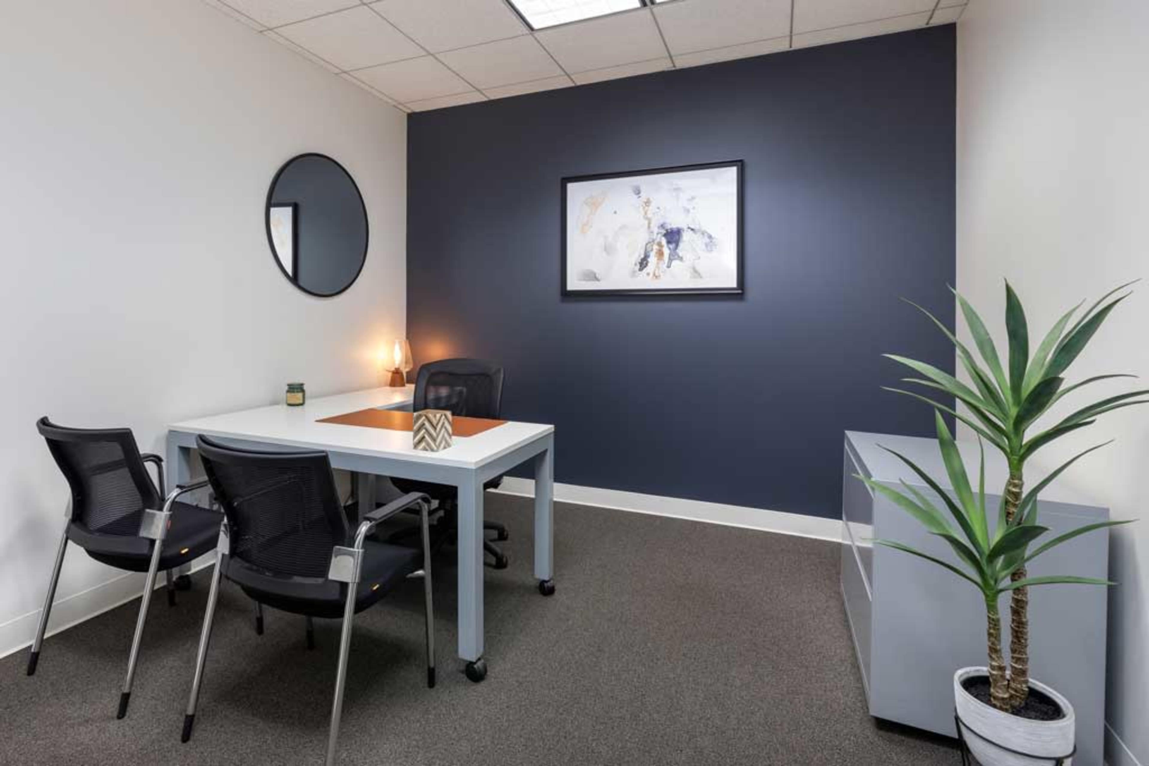 A modern office space featuring a round mirror, a desk with two chairs, a wall art piece, and a potted plant in the corner.