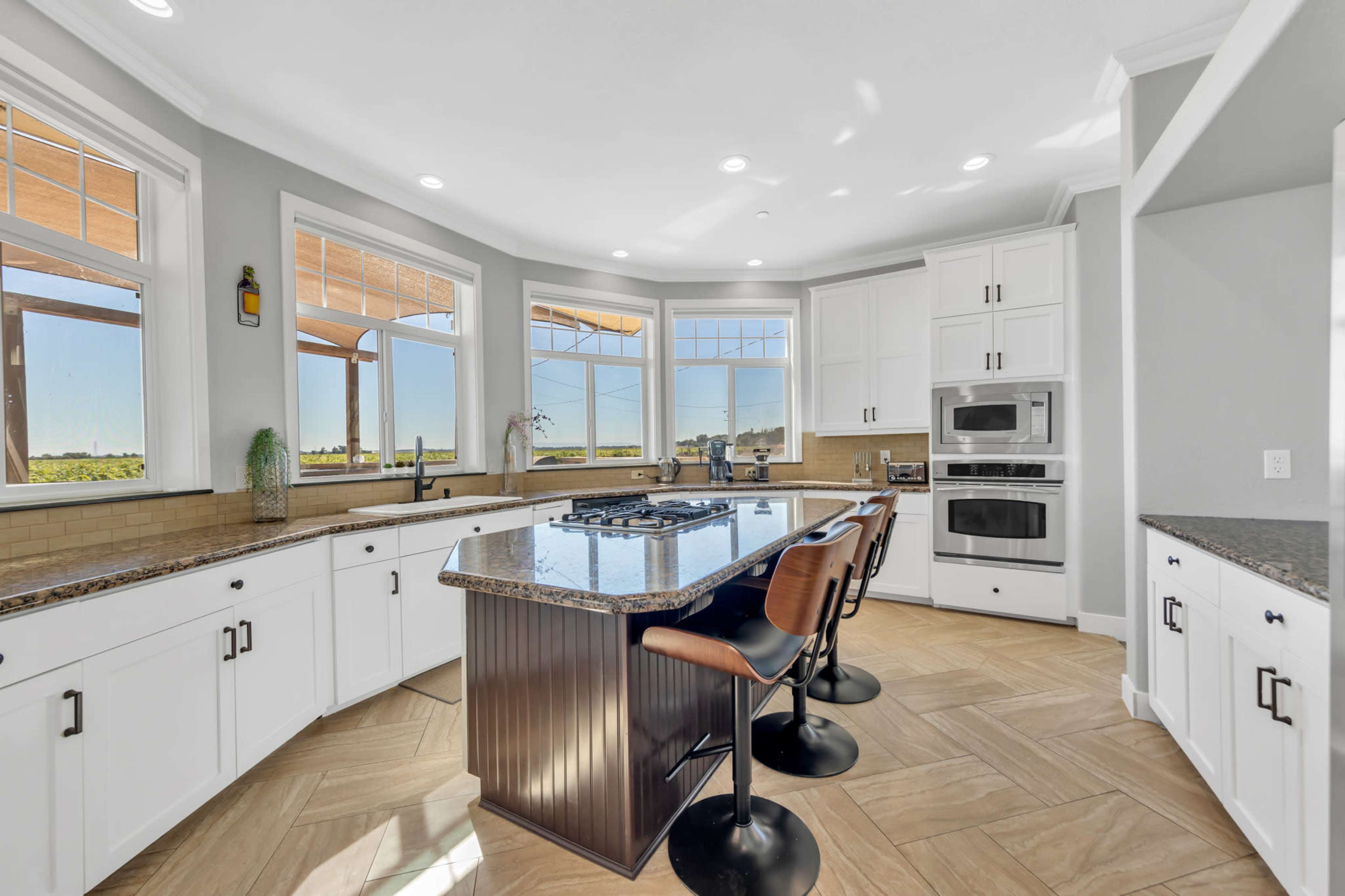 A modern kitchen features white cabinetry, a large island with seating, and windows that provide ample natural light and a view of the outdoors.
