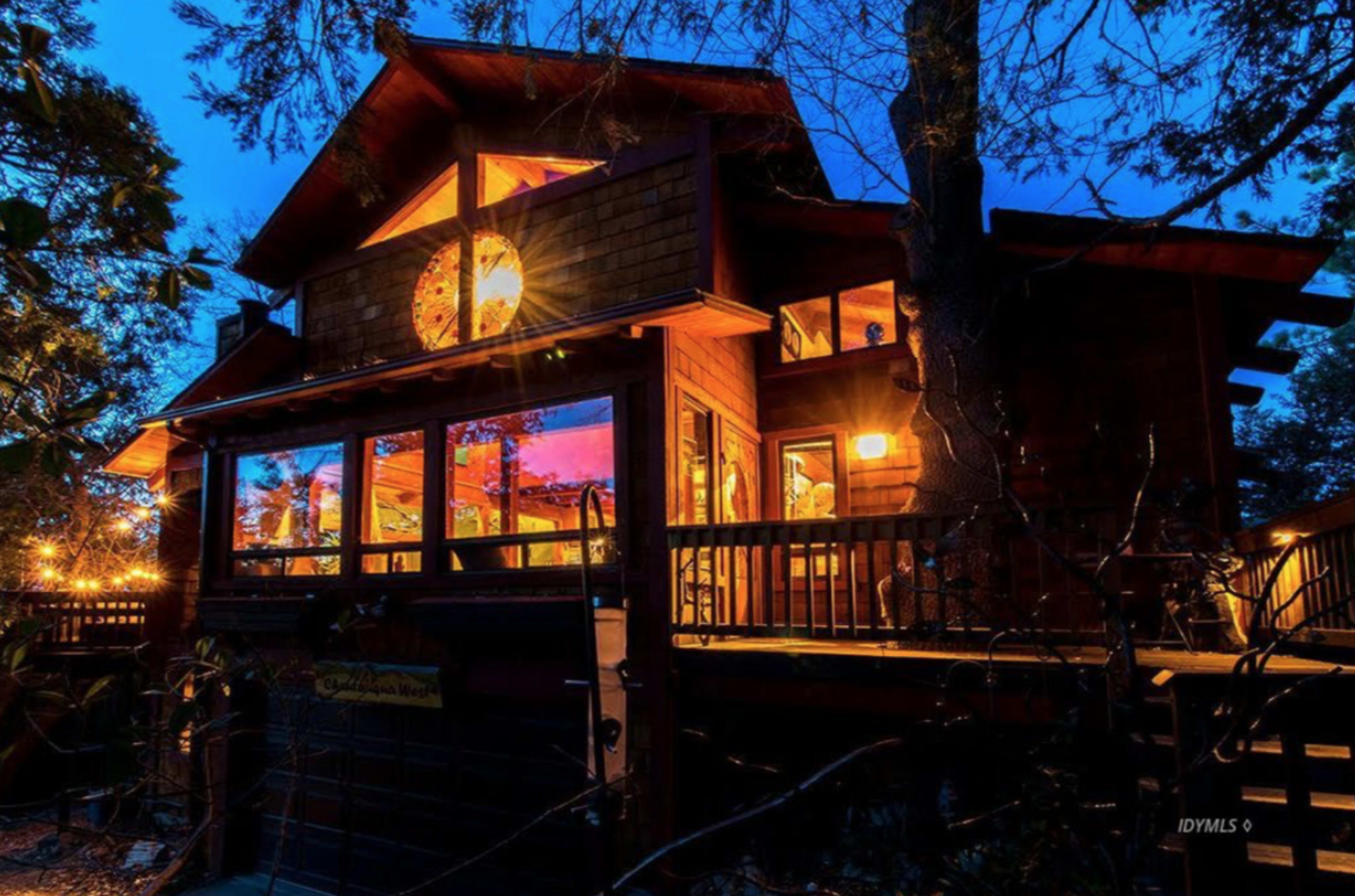 The image depicts a large wooden cabin with multiple windows illuminated in the dusk, surrounded by trees.