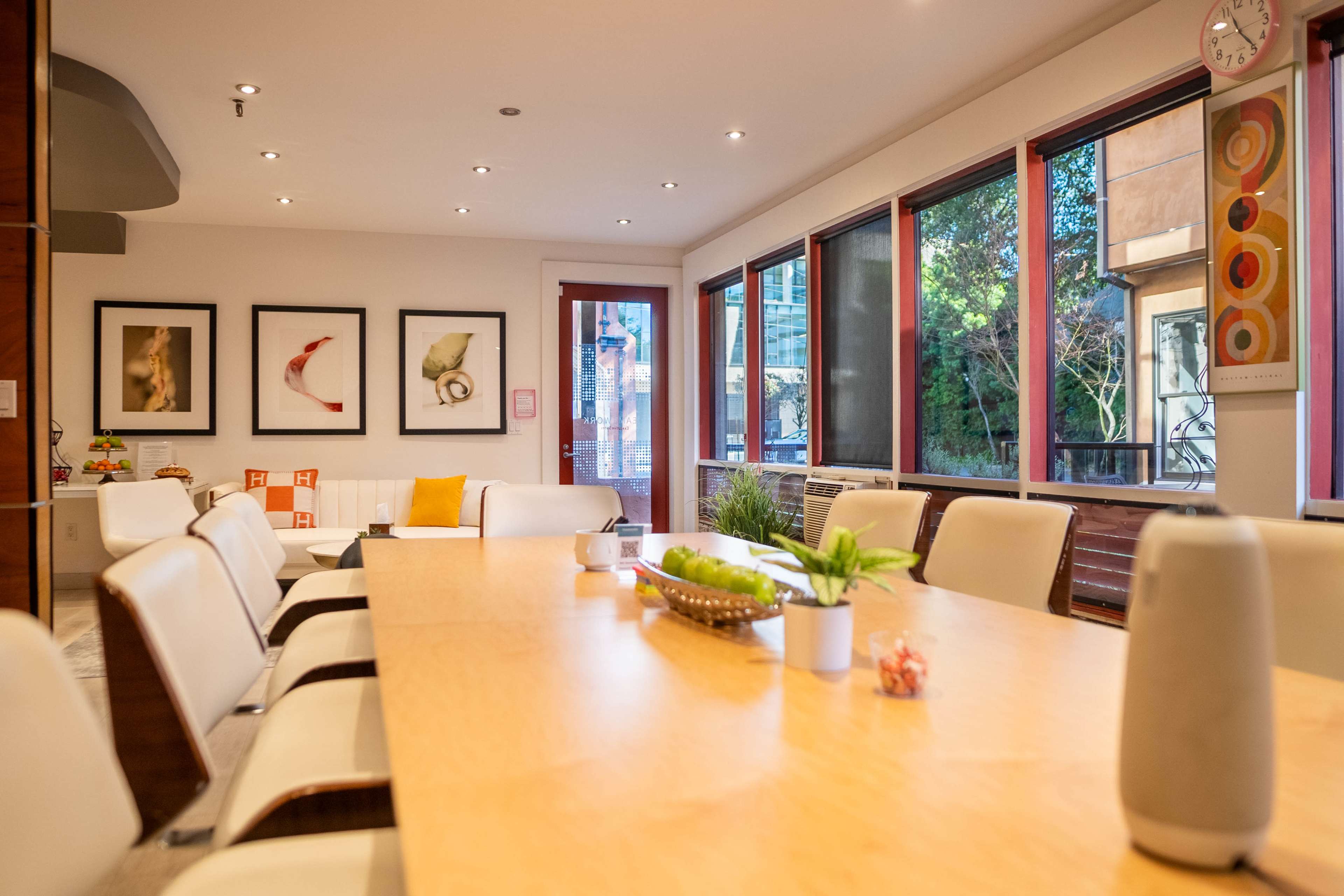A brightly lit conference room features a long wooden table surrounded by white chairs, with large windows displaying greenery outside.