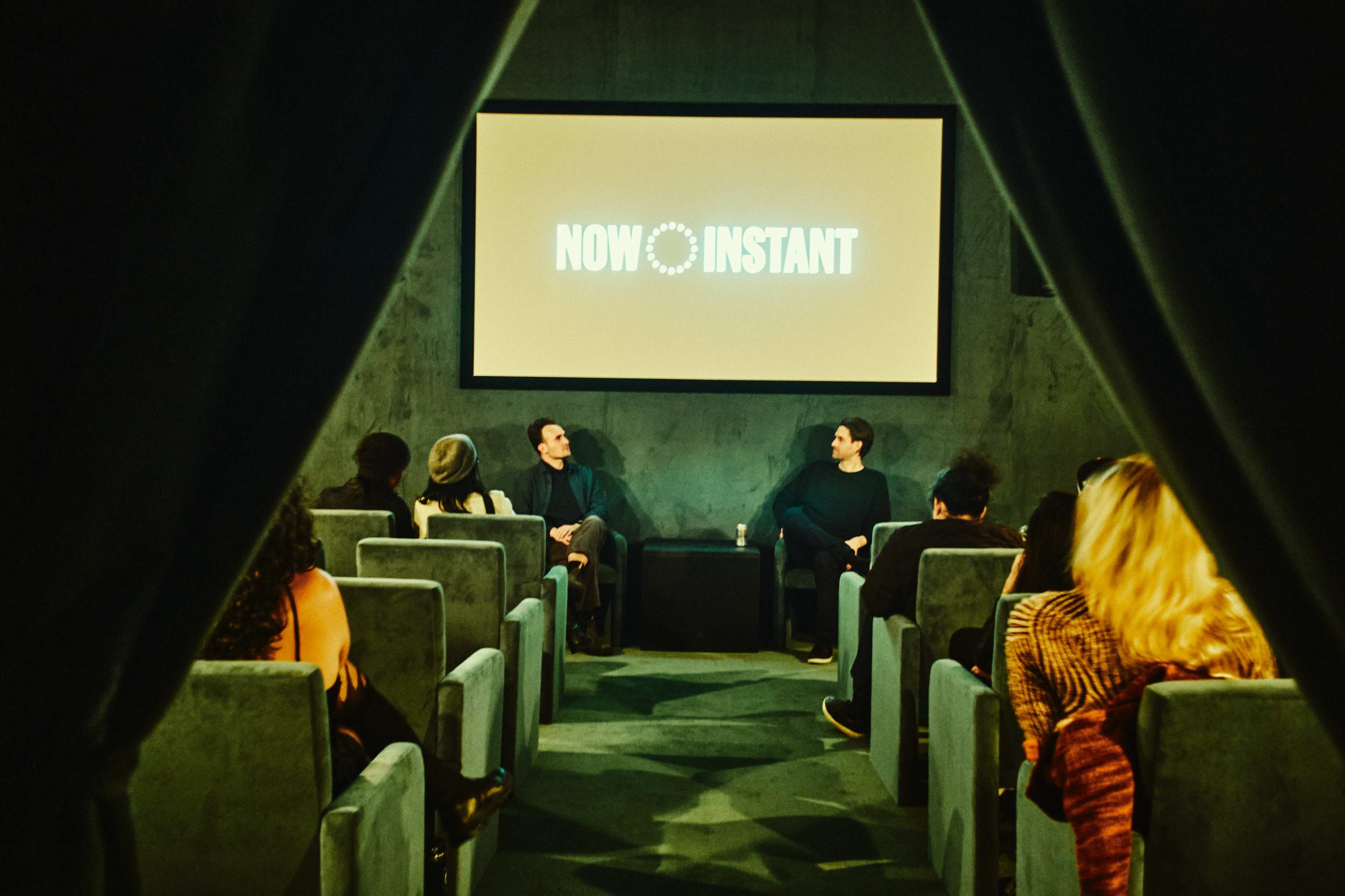 A small audience sits in a theater-like setting, facing a screen displaying the text "NOW INSTANT" while two speakers engage in conversation on stage.