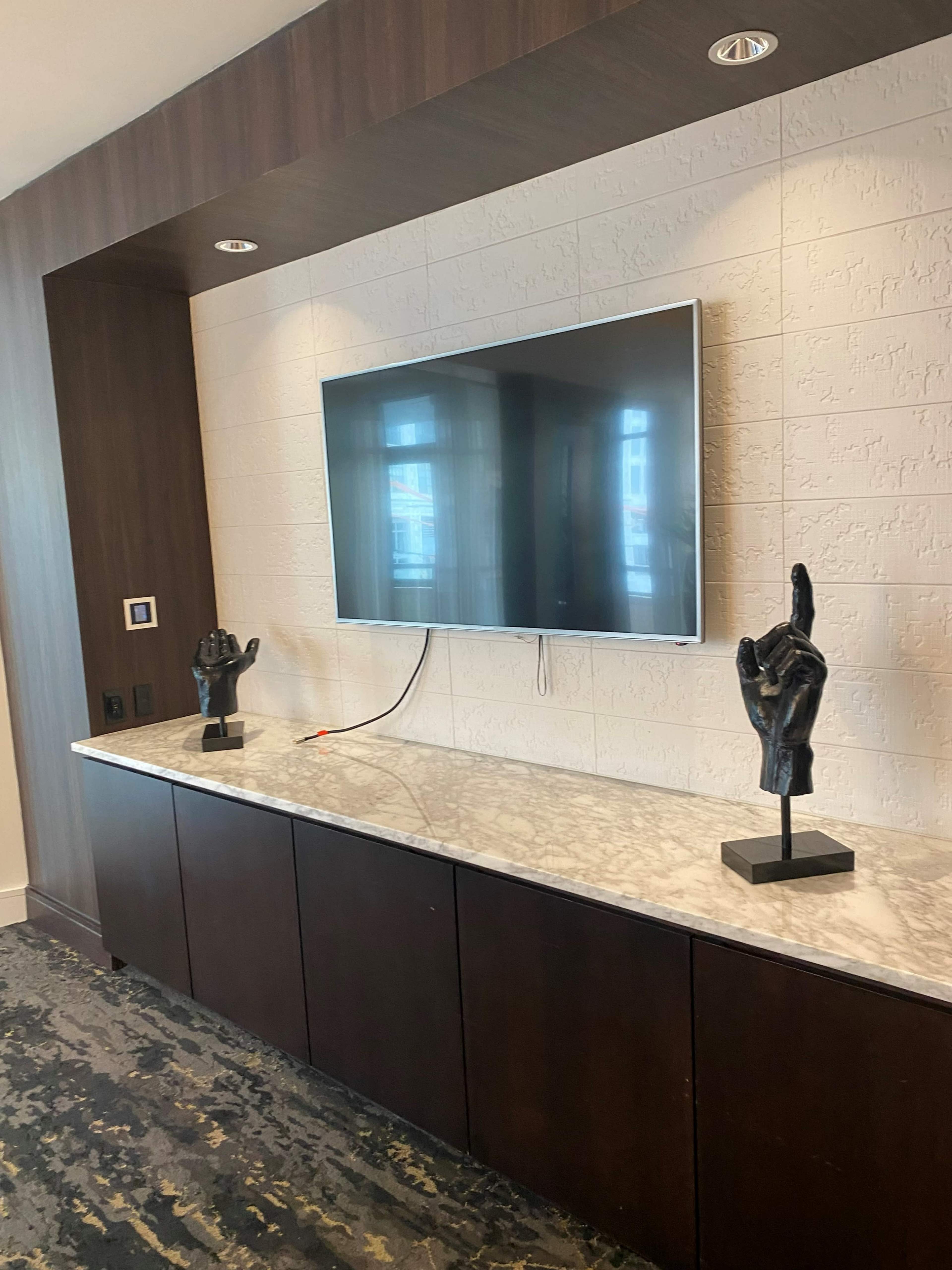 A wall-mounted television above a marble countertop with two decorative black hand sculptures on pedestals.