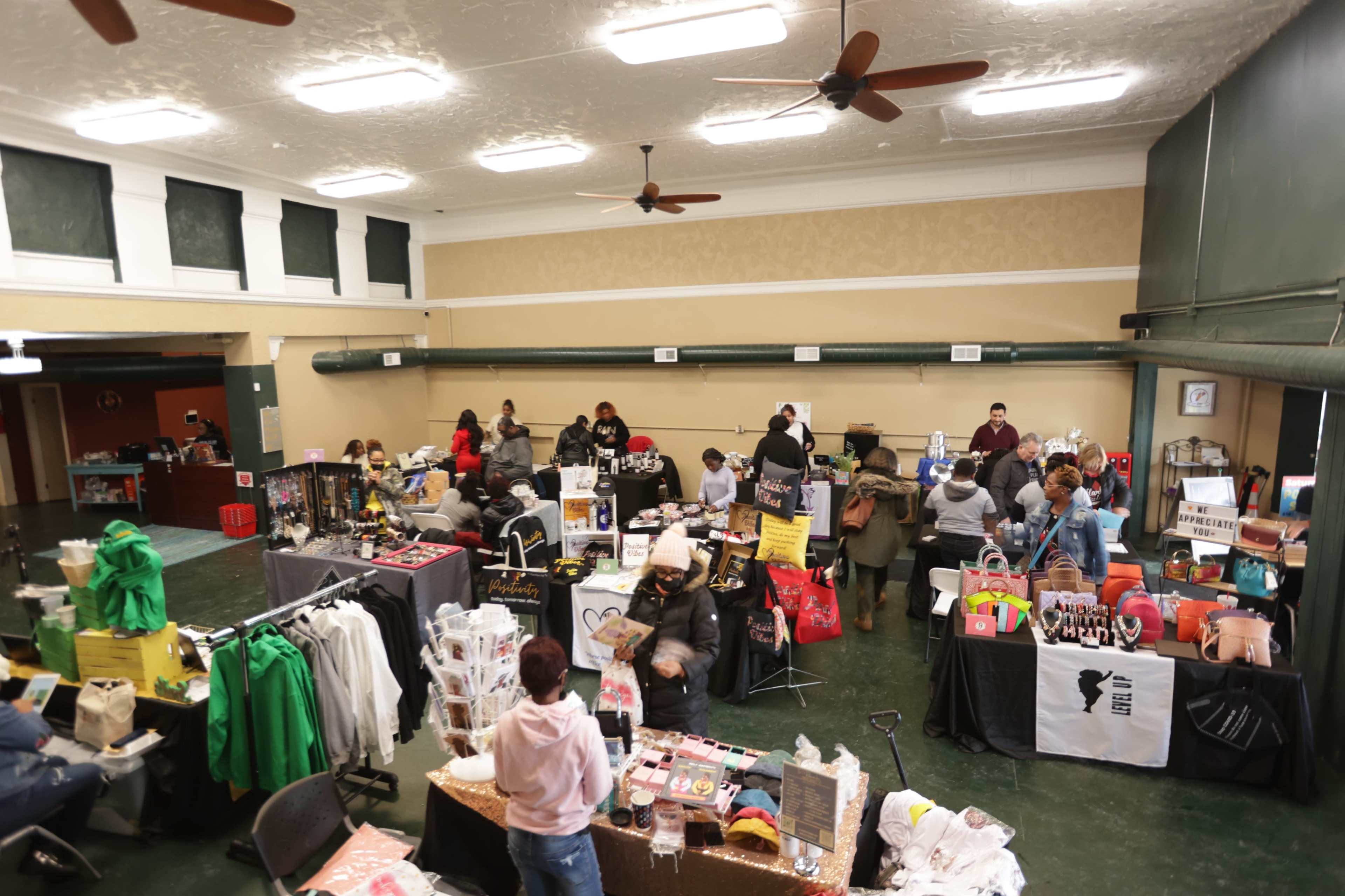 A large community space is set up for a marketplace, with several vendors displaying a variety of products while customers browse and socialize.