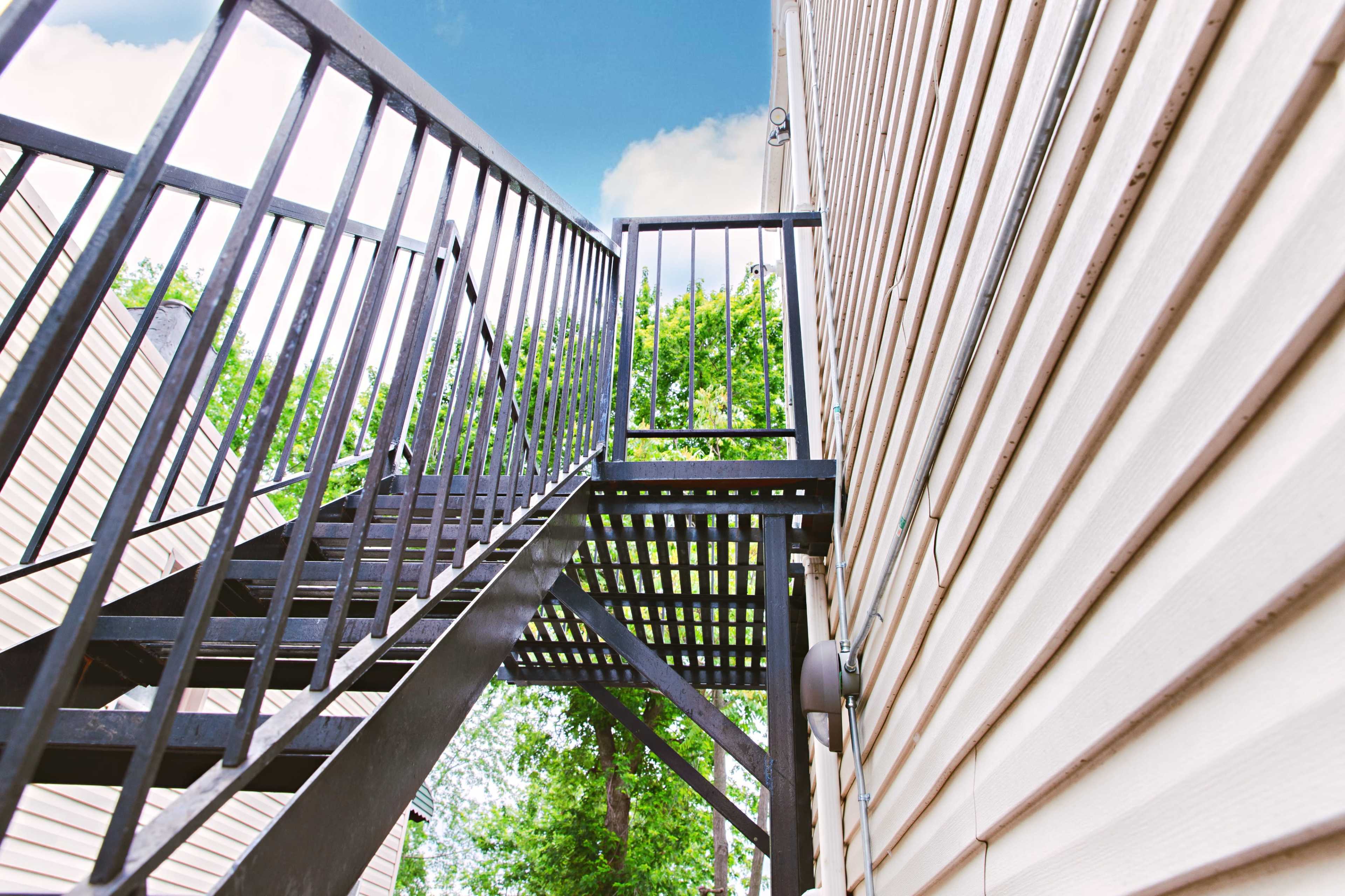 A metal outdoor staircase ascends beside a wooden building, framed by green trees and a blue sky.