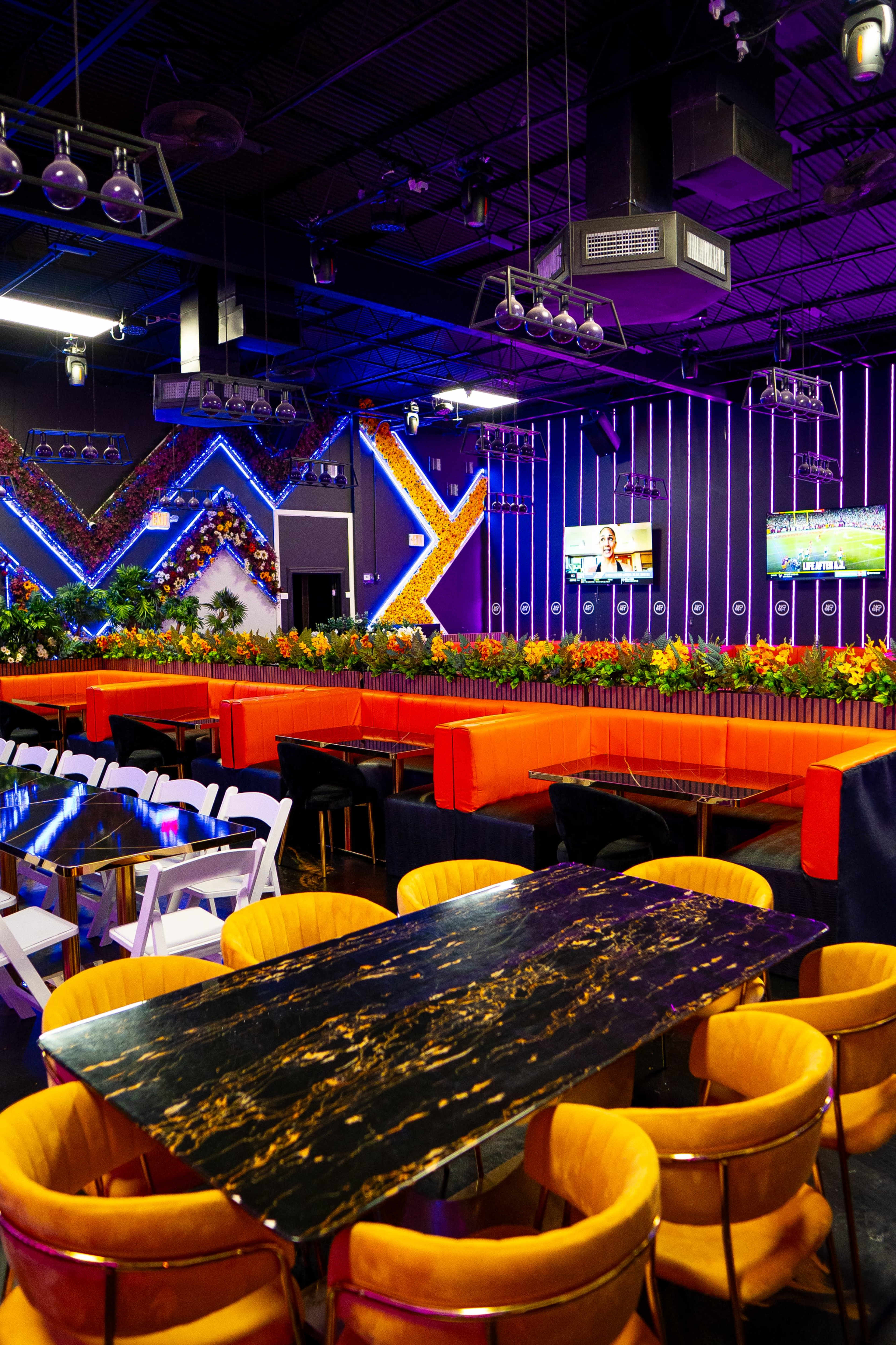 A modern venue features vibrant seating arrangements with colorful accents and multiple screens displaying sports, surrounded by lush decorations.