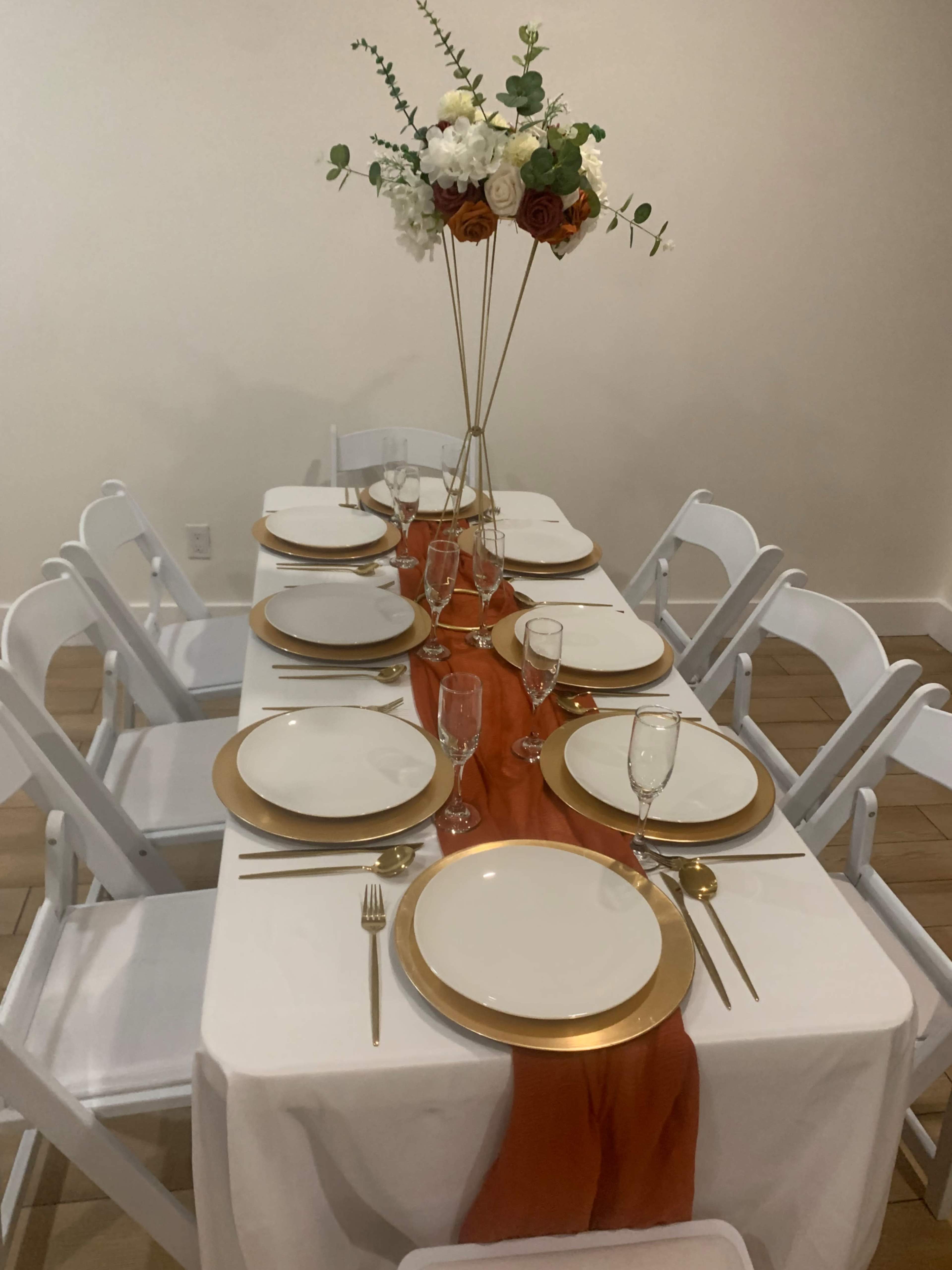 A dining table is set with white plates, gold chargers, glassware, and a centerpiece of flowers, arranged on a white tablecloth with a rust-colored runner.