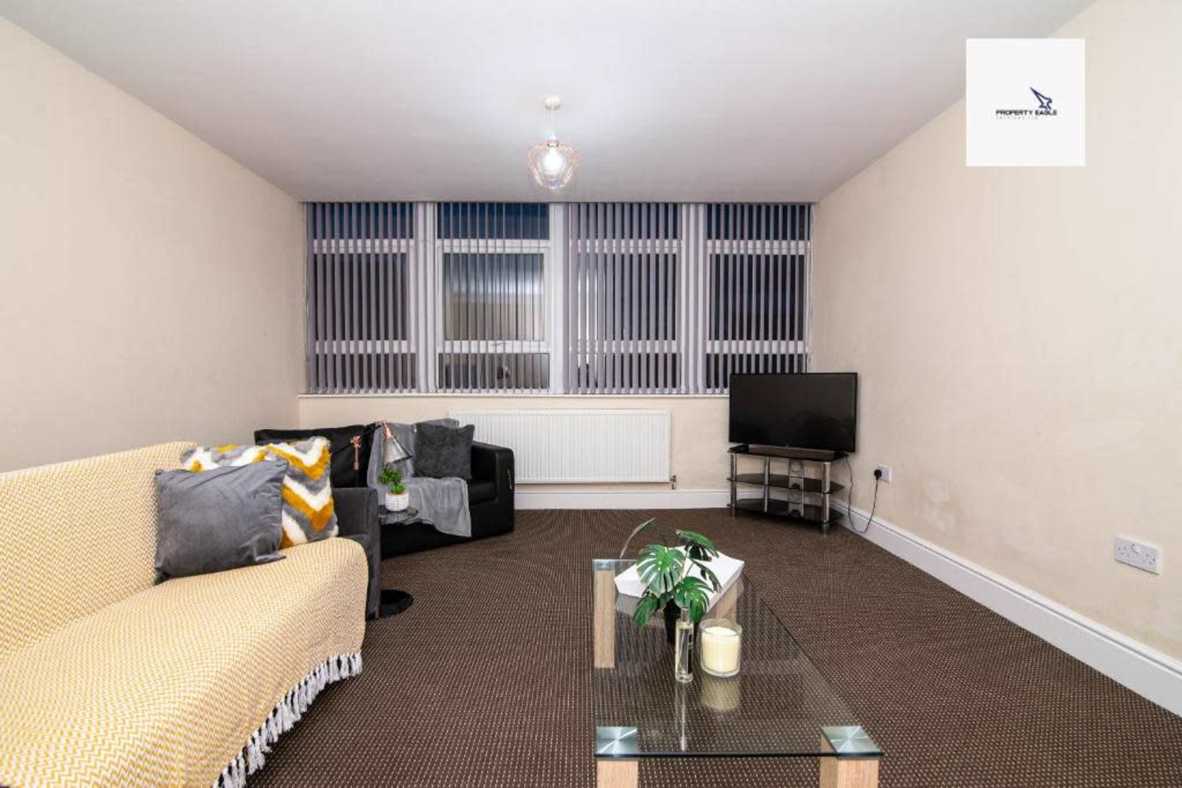 A living room features a yellow and white couch, a glass coffee table with a plant, a television on a stand, and large windows with vertical blinds.
