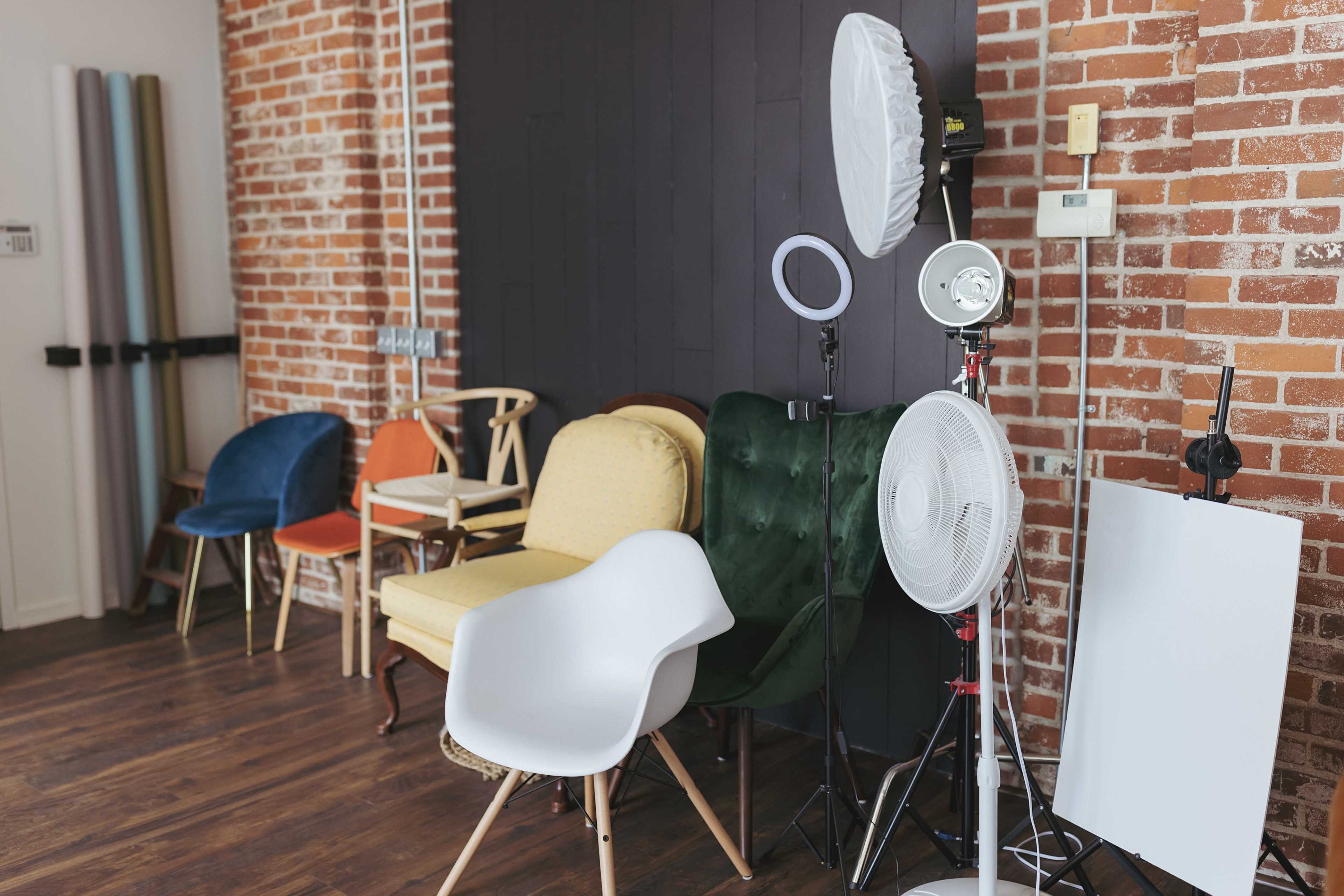 A variety of chairs and photography equipment arranged against a brick wall in a studio setting.