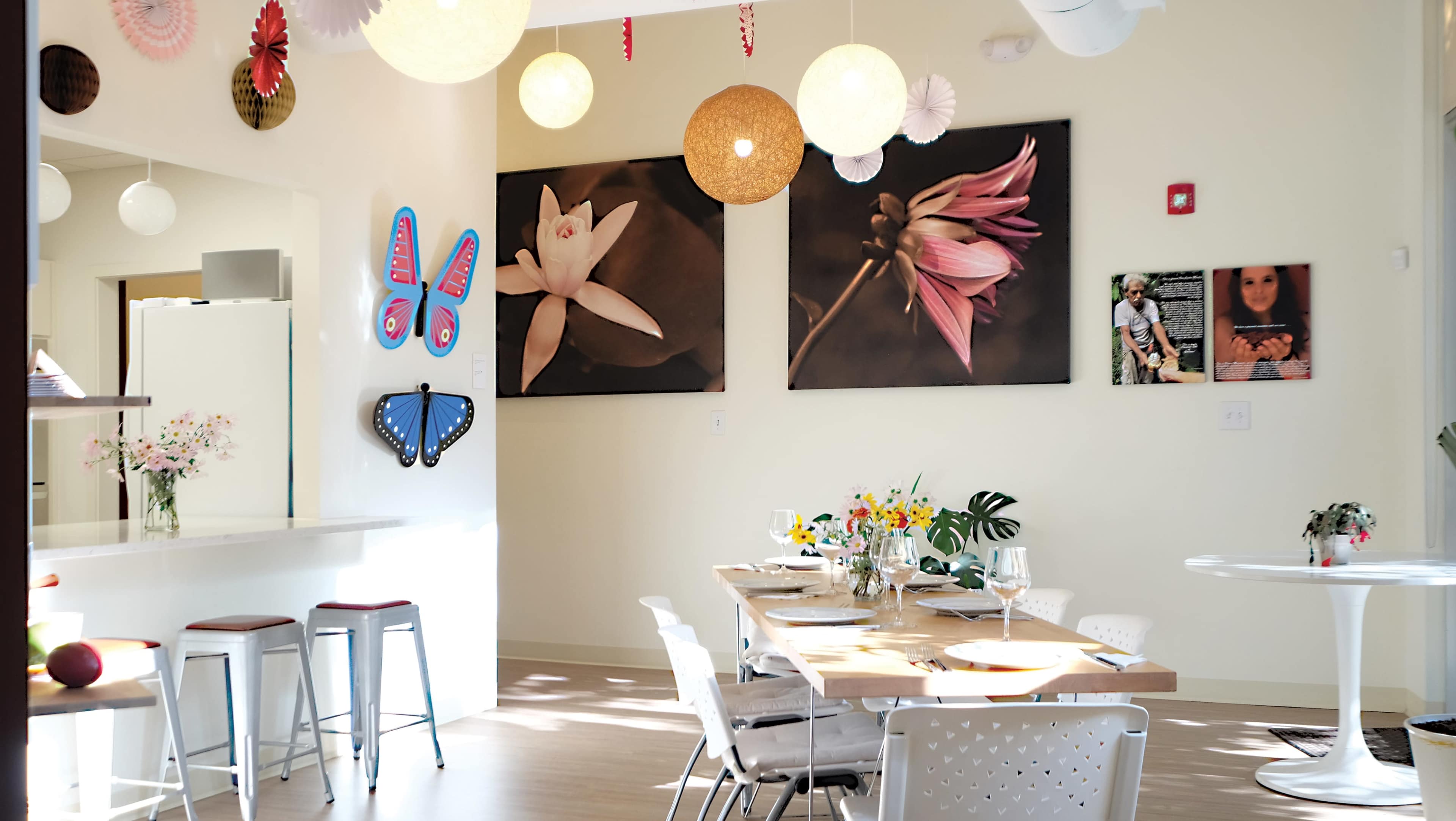 A bright dining area features a wooden table set for a meal, surrounded by white chairs, with colorful wall decorations and large floral photographs.