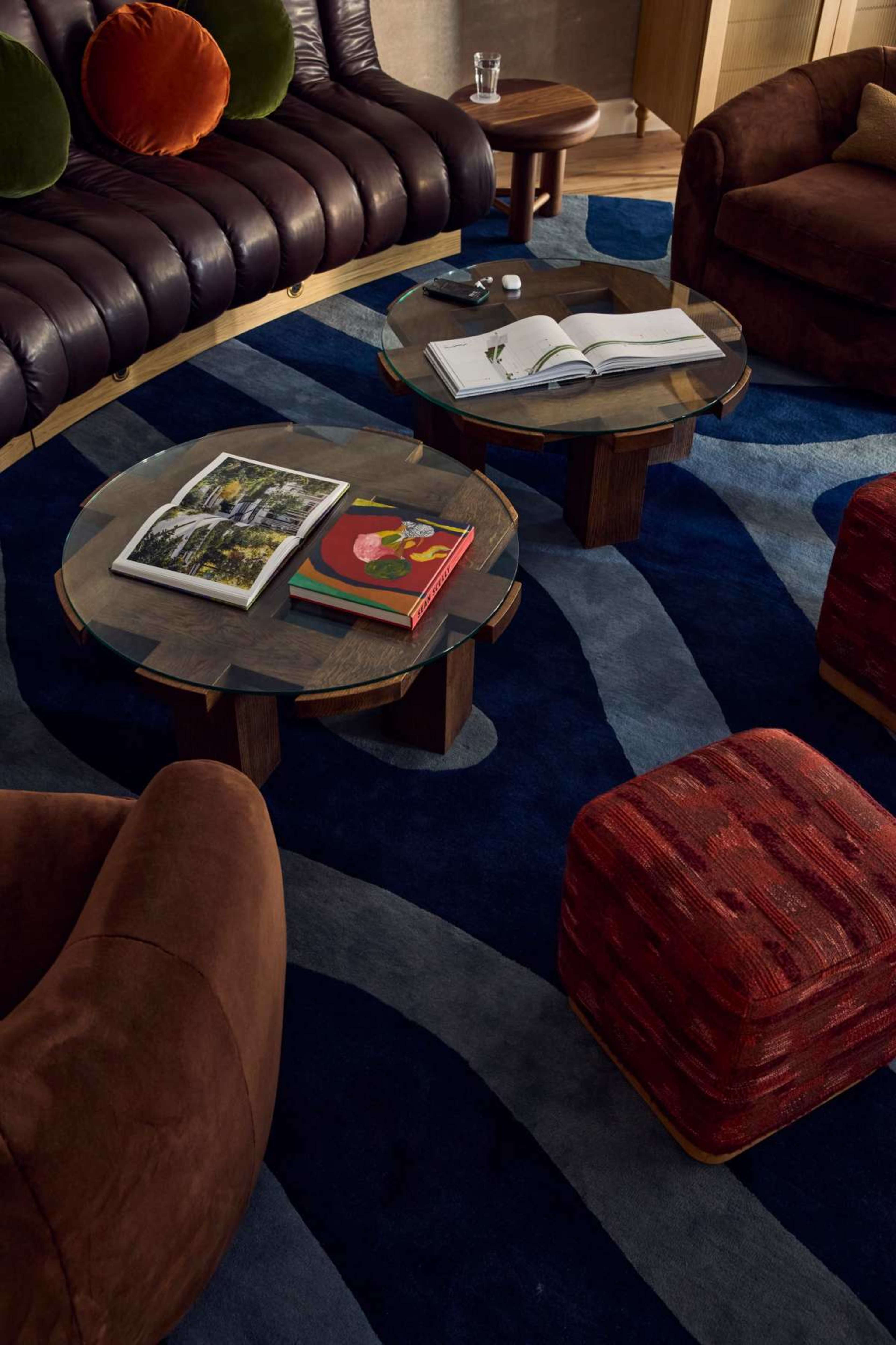 The image features a modern living room with curved, brown furniture, two round wooden coffee tables, and a large, patterned area rug.
