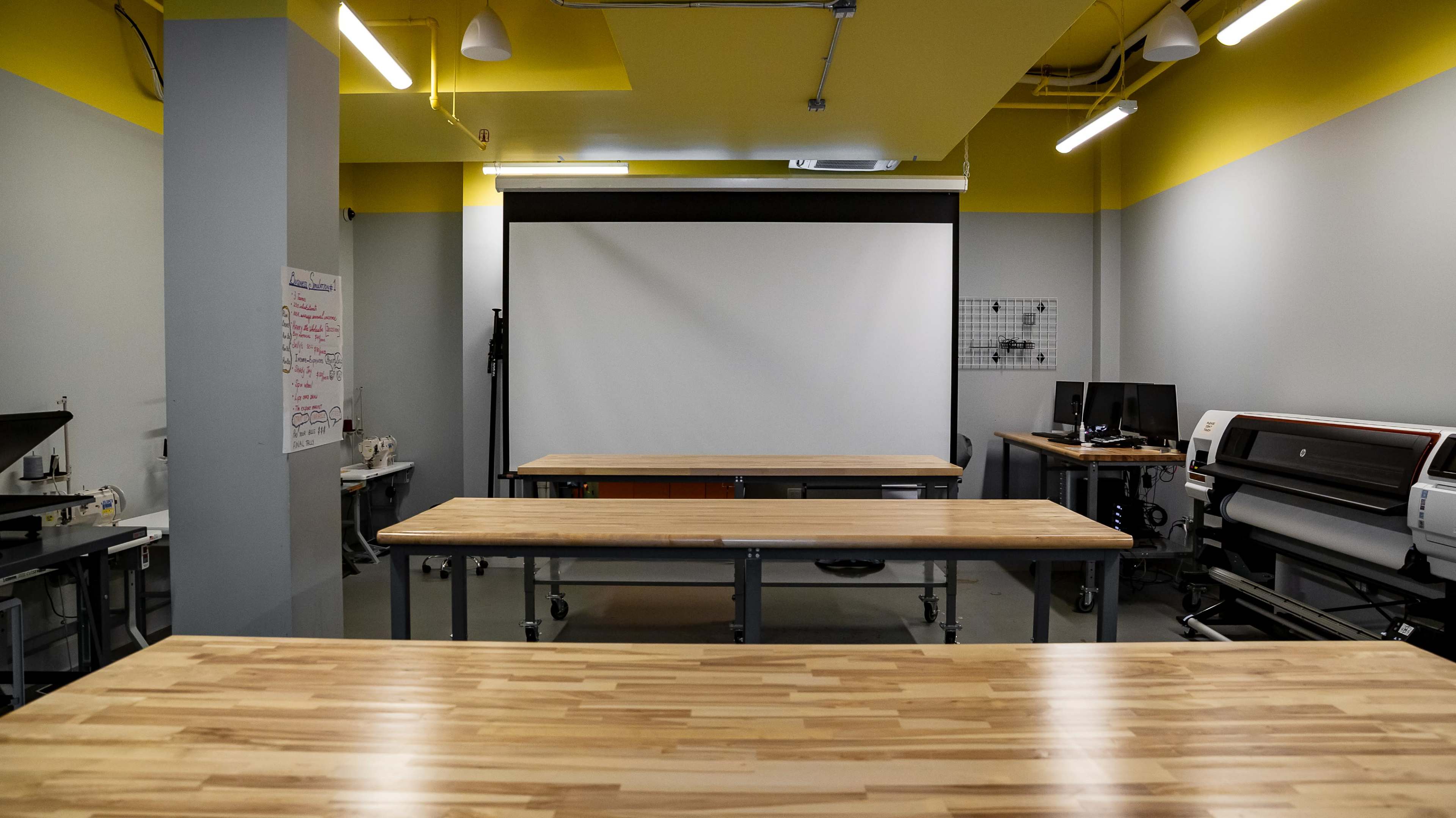 The image shows a spacious workshop or classroom area with wooden tables, a large projection screen, and various equipment along the walls.
