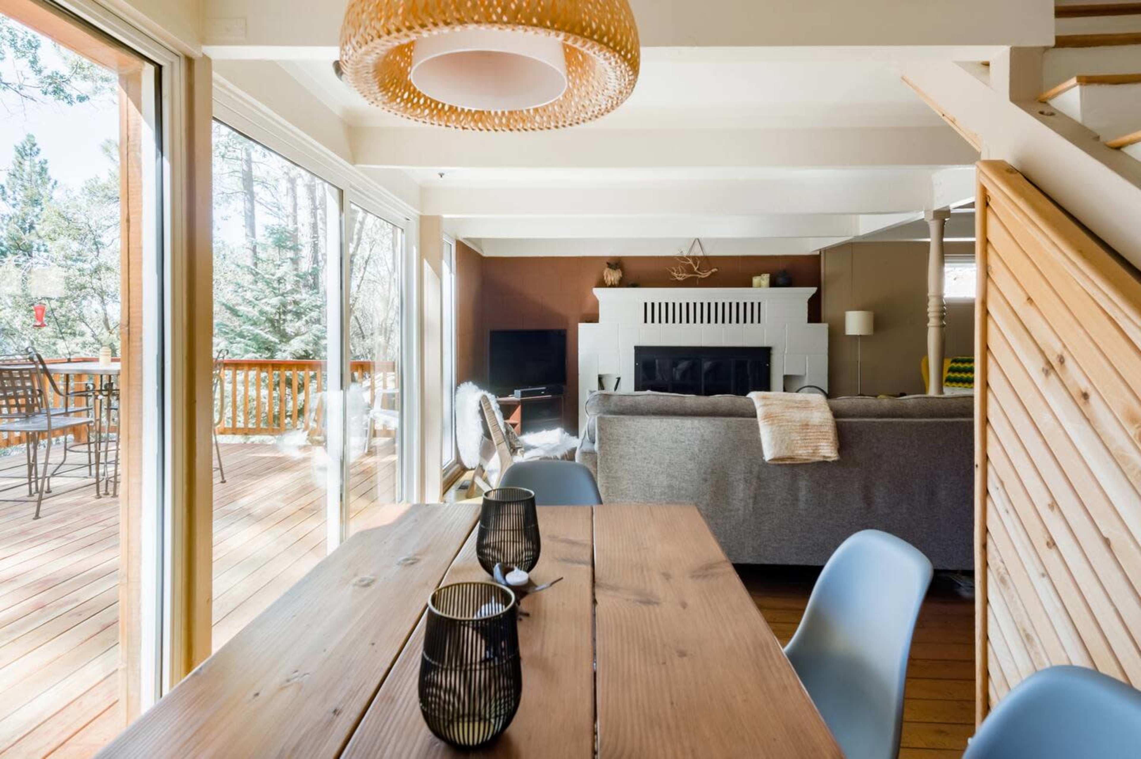 A dining area with a wooden table and two black glasses, adjacent to a living room featuring a gray sofa and a fireplace, with large glass doors opening to a deck.