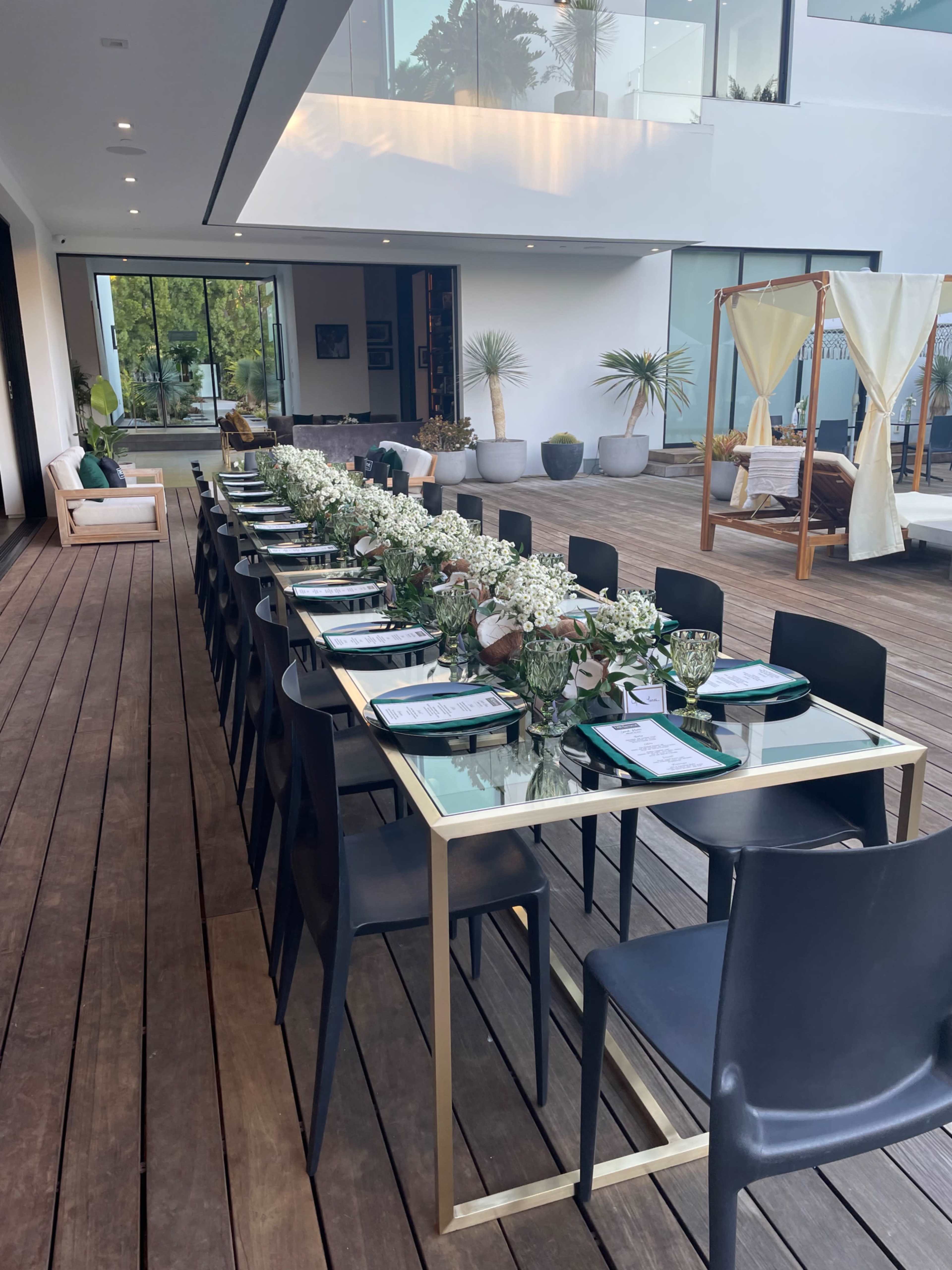 A long, elegantly set dining table with floral centerpieces is positioned on a wooden deck adjacent to a modern home.