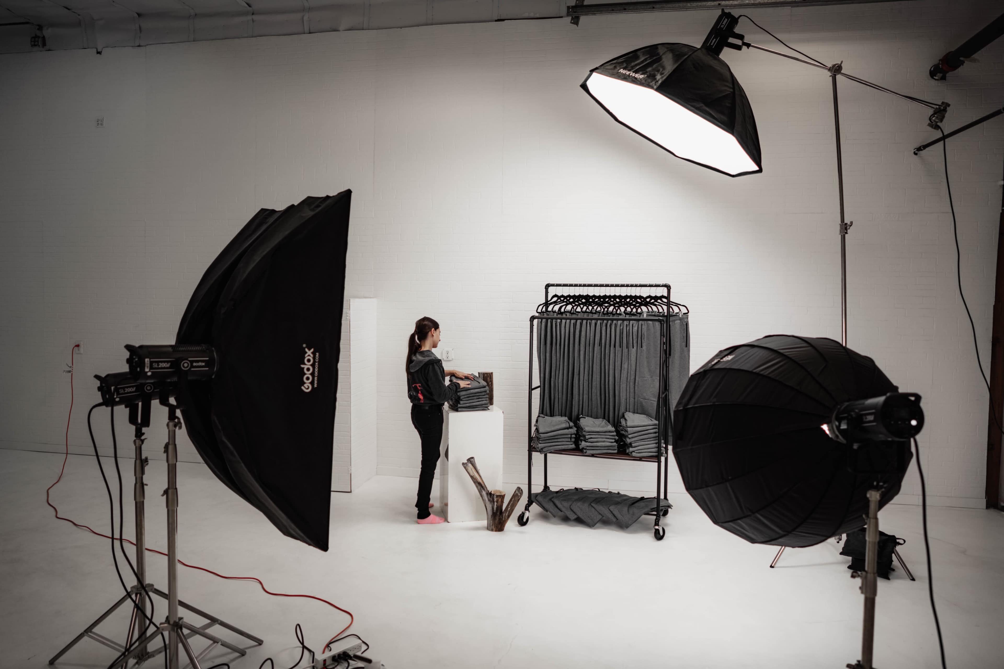 A woman organizes clothing on a rack in a photography studio equipped with several lighting setups.