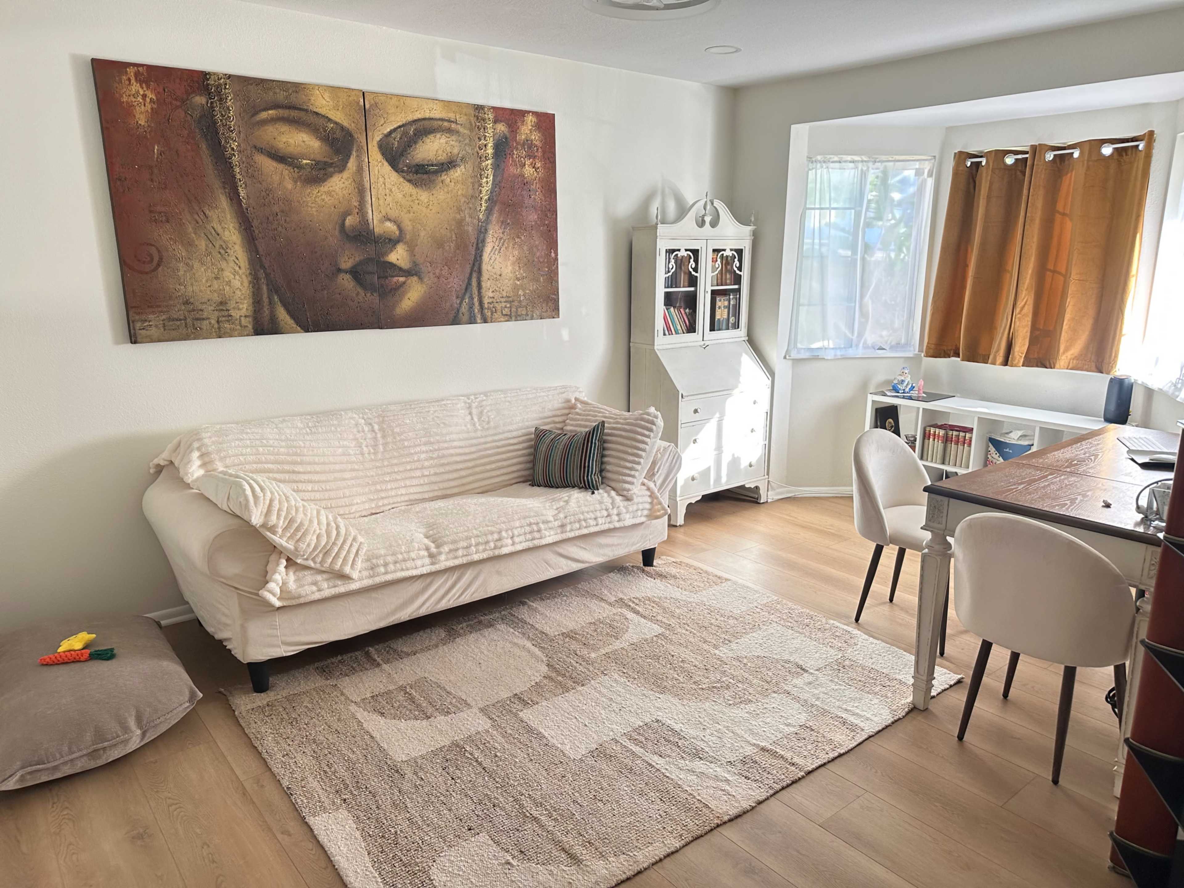 The image depicts a living room featuring a sofa, a decorative wall art of a face, a dining table with chairs, and natural light coming through a window with curtains.
