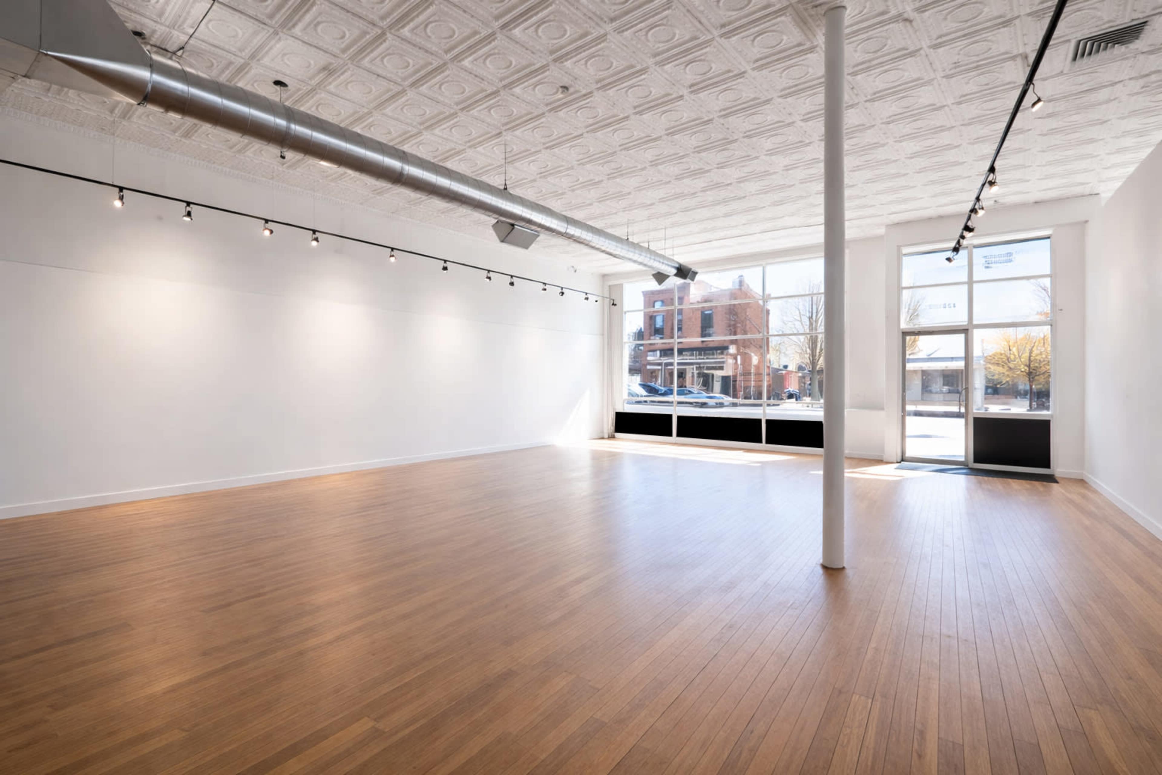 The image shows a spacious, empty room with wooden flooring and large windows that provide natural light.