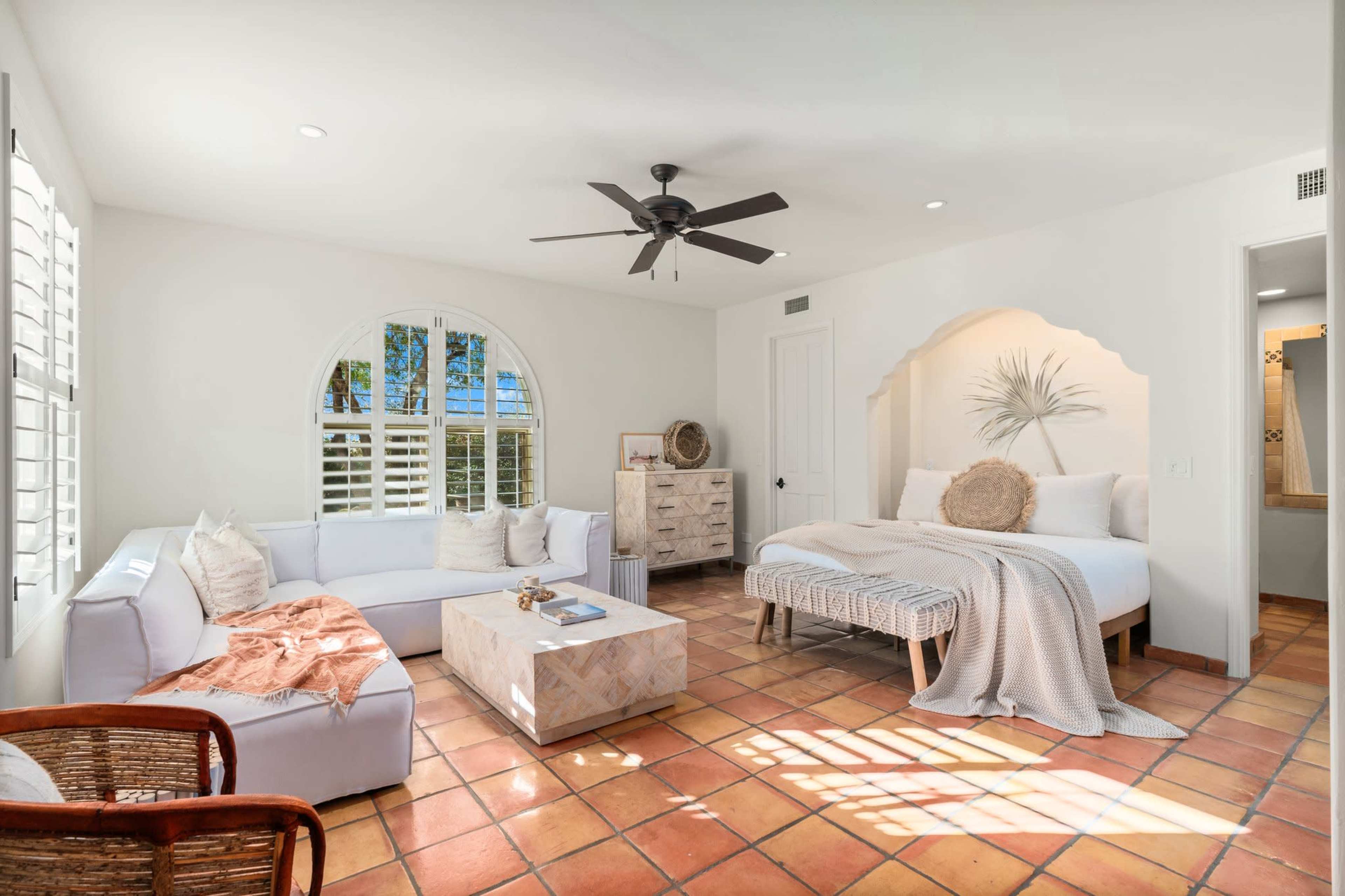 A bright, well-lit room featuring a white sofa, a coffee table, and a bed with a textured blanket against a wall with a decorative arch.