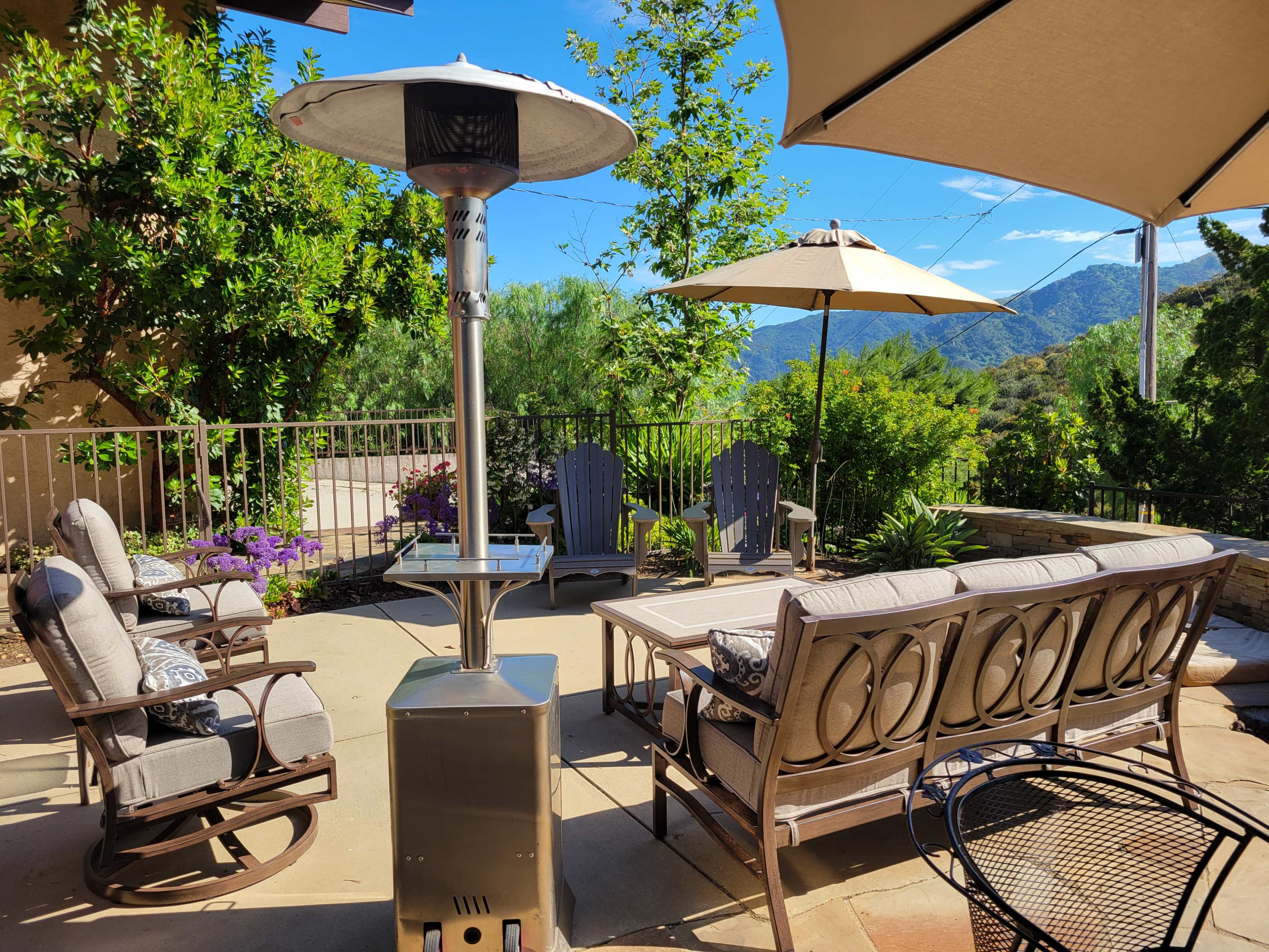 A patio area features comfortable seating, a patio heater, and two umbrellas, surrounded by greenery and mountain views.