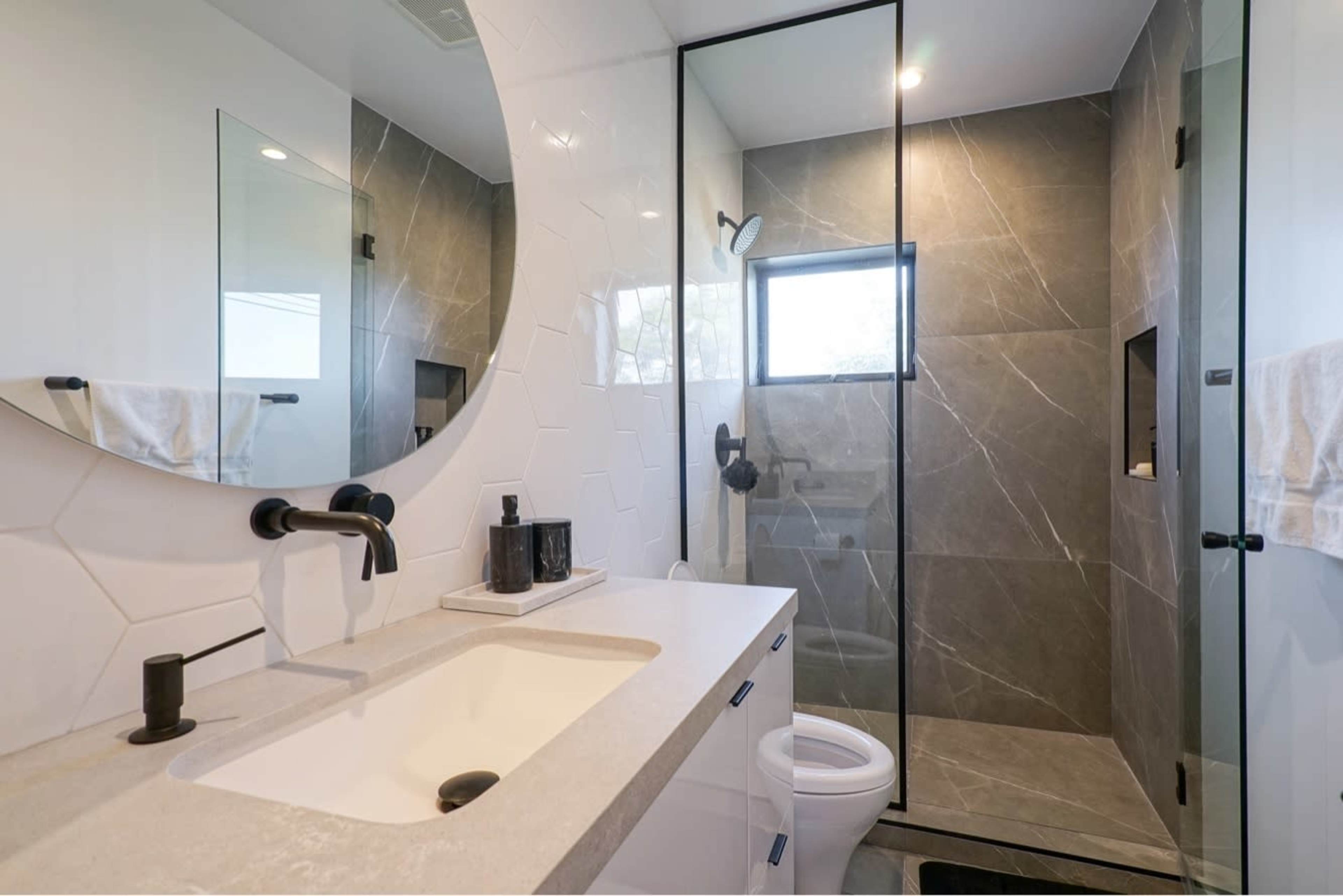 The image shows a modern bathroom with a circular mirror, white countertop, and a glass-enclosed shower featuring gray tiles.
