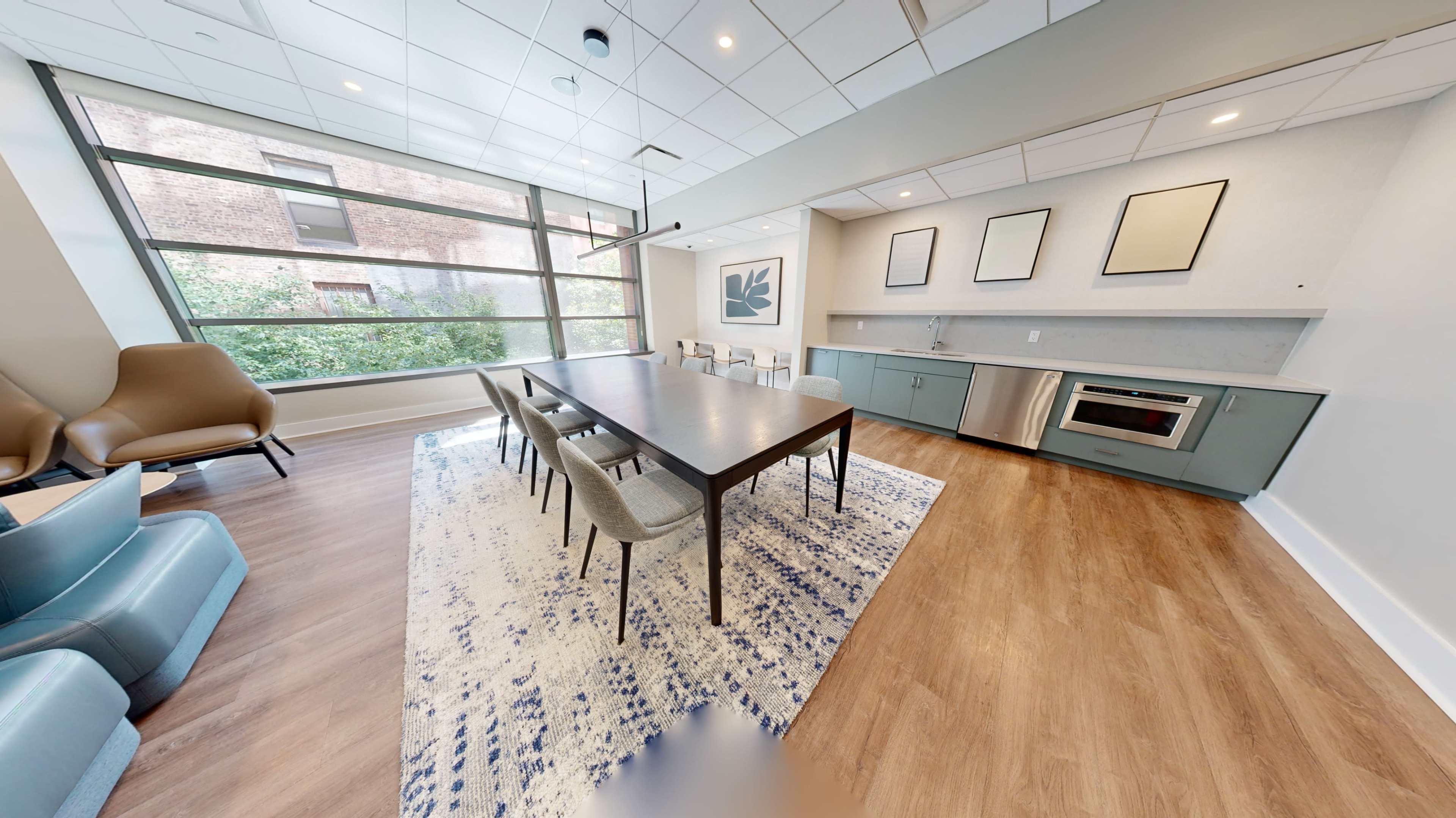 The image shows a modern meeting room with a long table and chairs, alongside a kitchenette area featuring appliances and large windows letting in natural light.