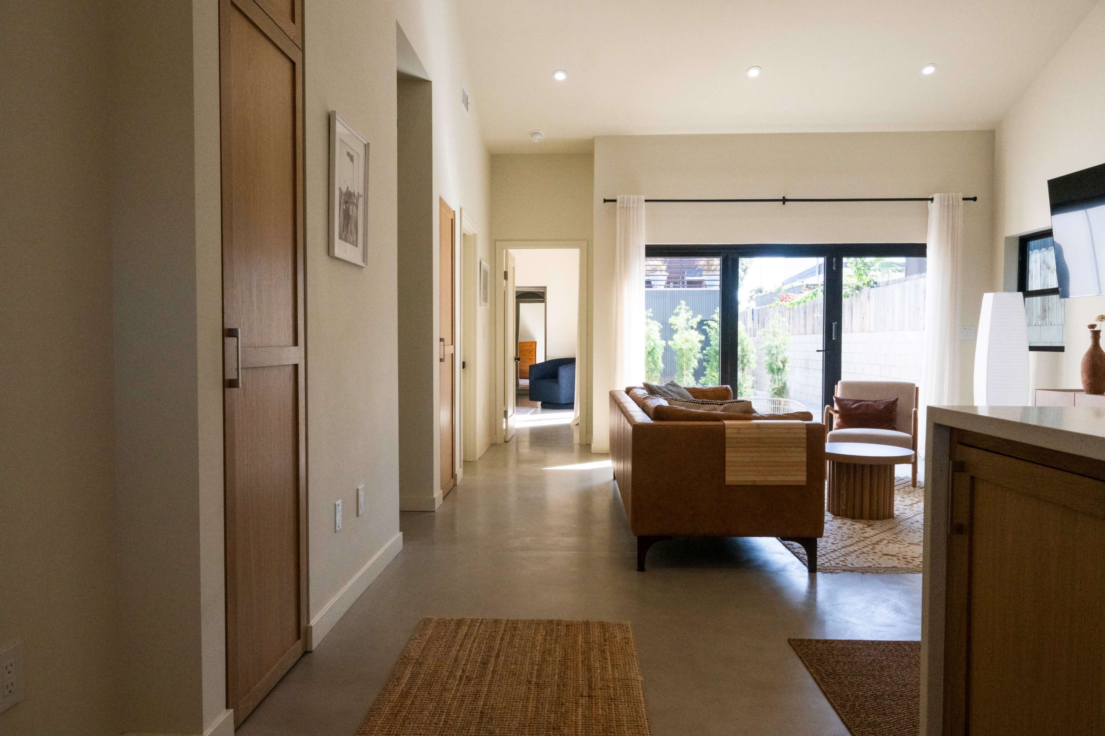 The image shows a modern interior hallway leading to a living area with a sofa and large windows, featuring light-colored walls and a polished concrete floor.