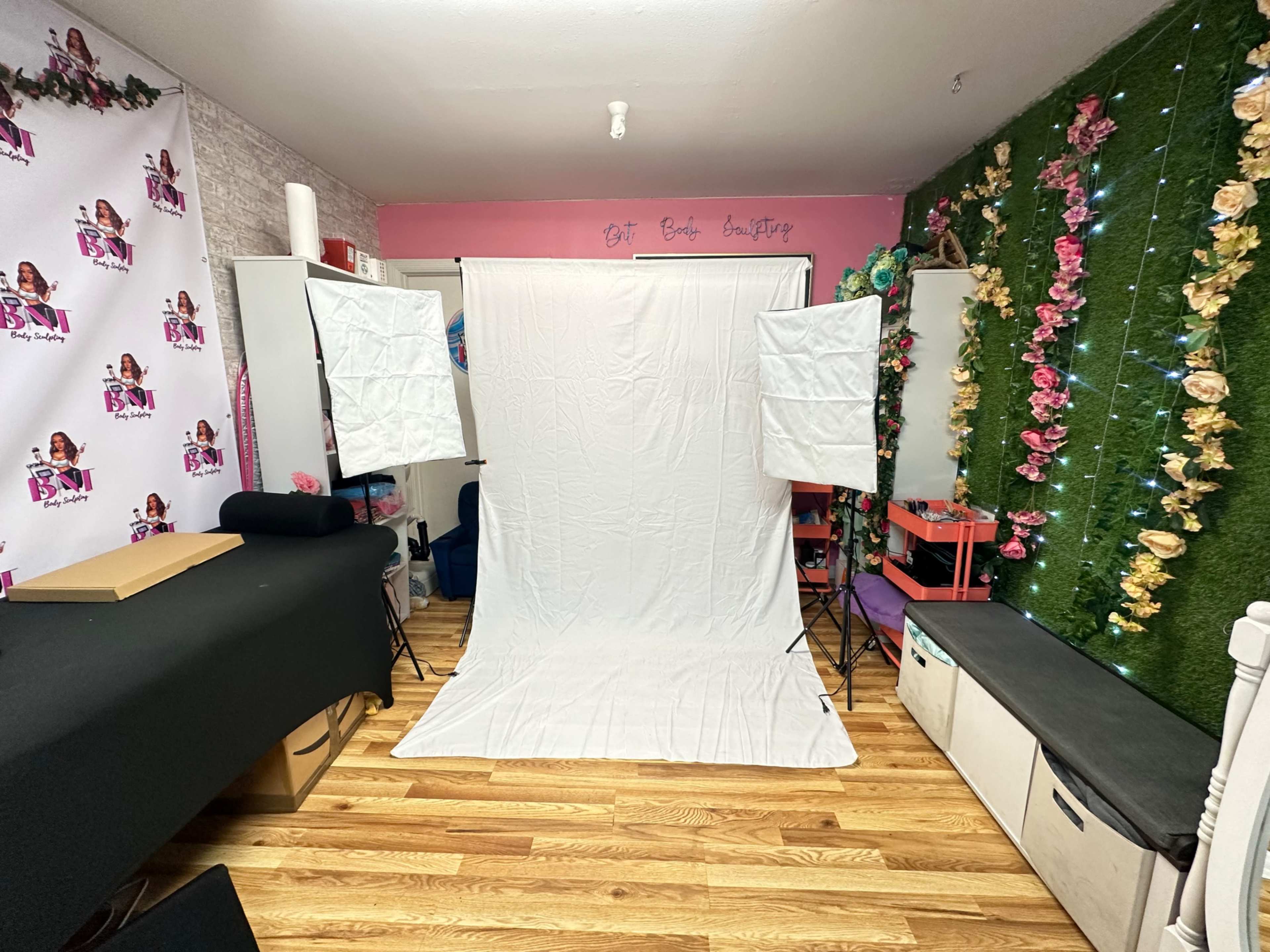 The image shows a studio space featuring a white backdrop, two lighting stands, and decorative floral elements on the walls.