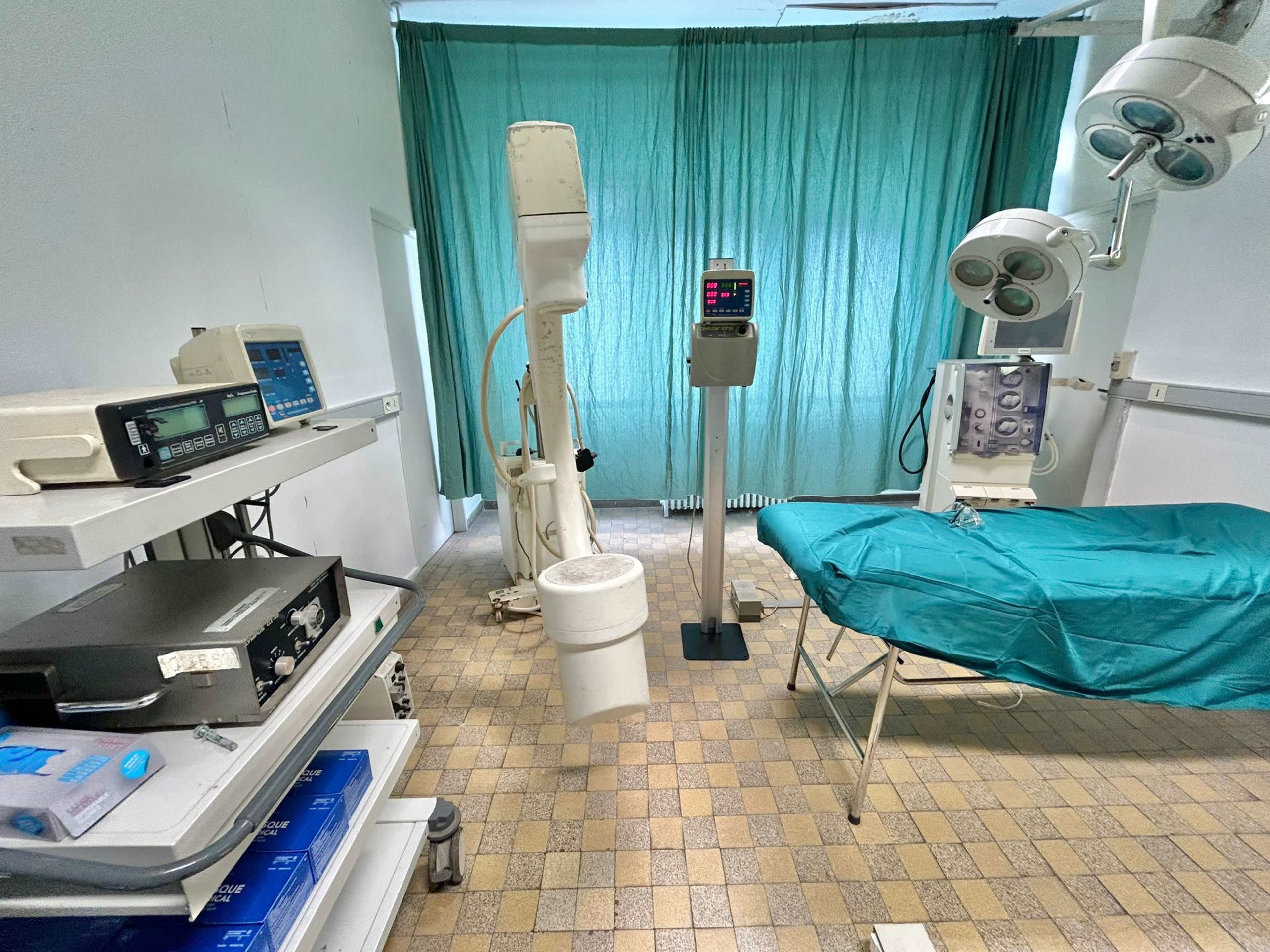 A medical examination room featuring a bed covered with a teal sheet, various medical equipment, and green curtains.