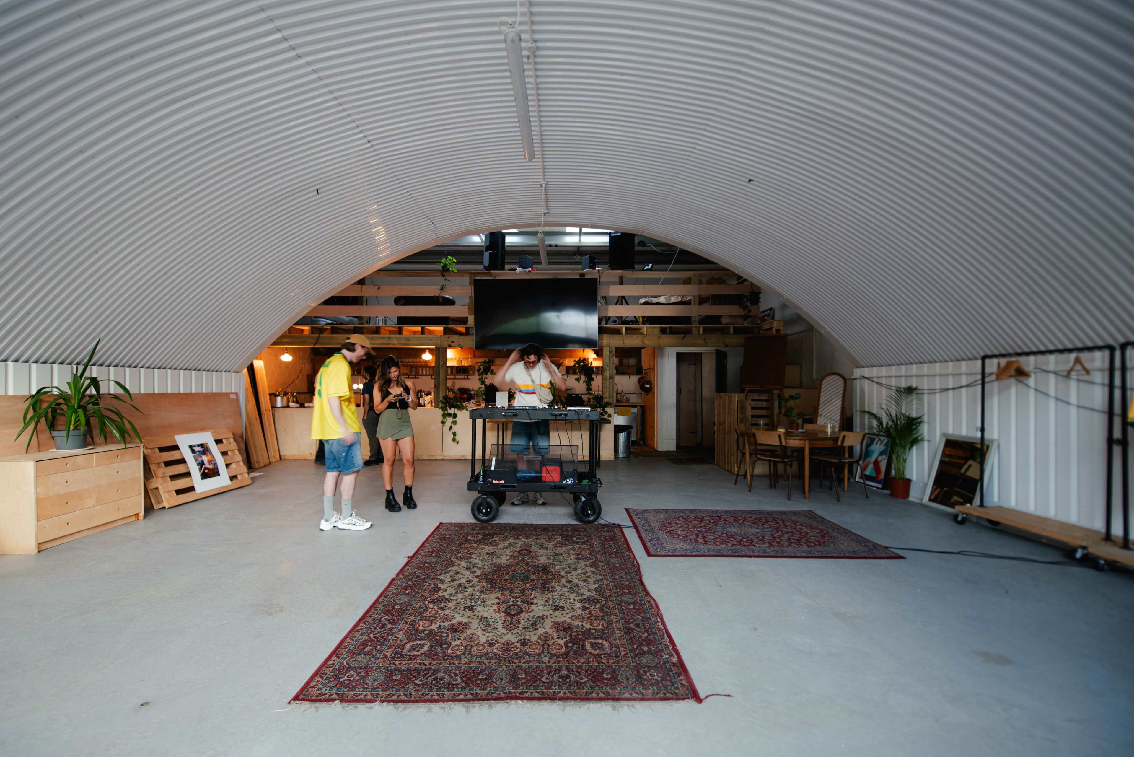 Two individuals stand near a cart in a spacious, arch-roofed interior with wooden furniture and plants, while another person appears to be preparing something on the cart.