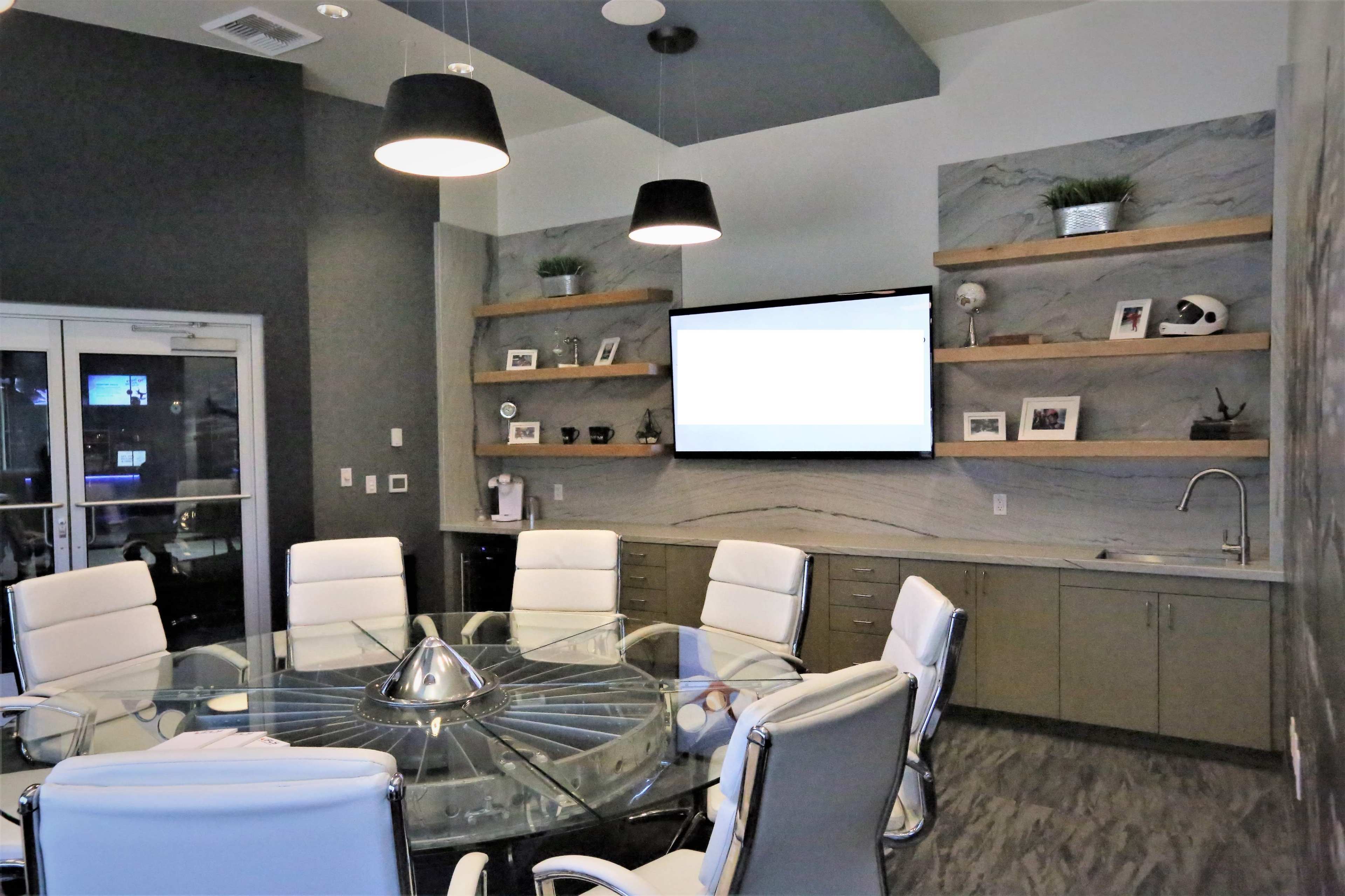 The image shows a modern conference room featuring a circular glass table surrounded by white leather chairs, with wall-mounted shelves displaying decorative items and a large screen at the front.