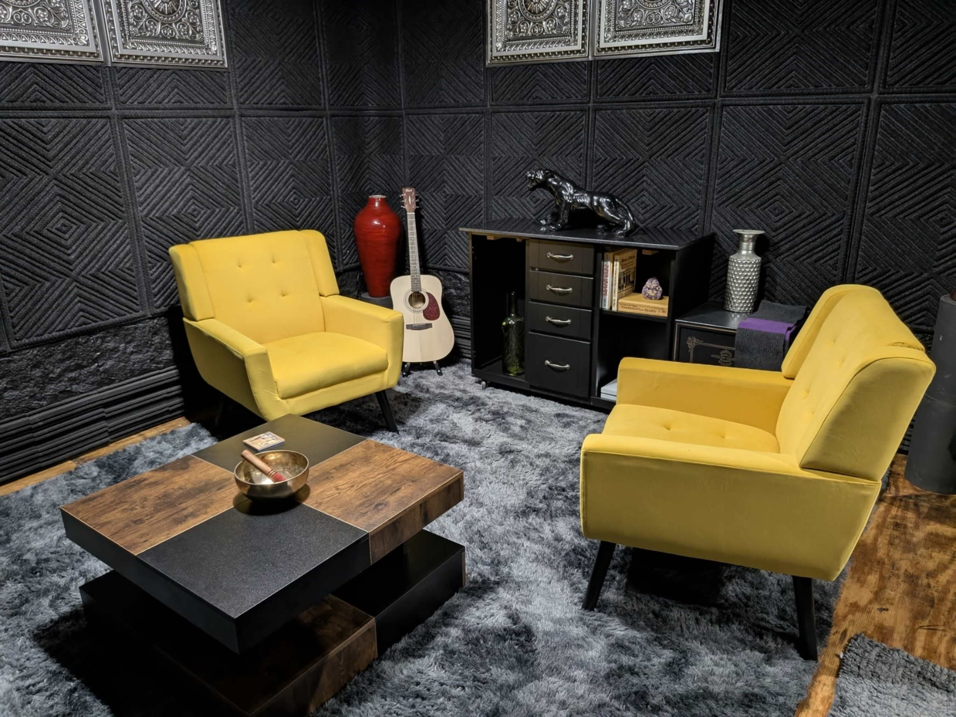 The image shows a cozy room with two yellow chairs, a wooden coffee table, and various decorative items, including an acoustic guitar and a black cabinet.