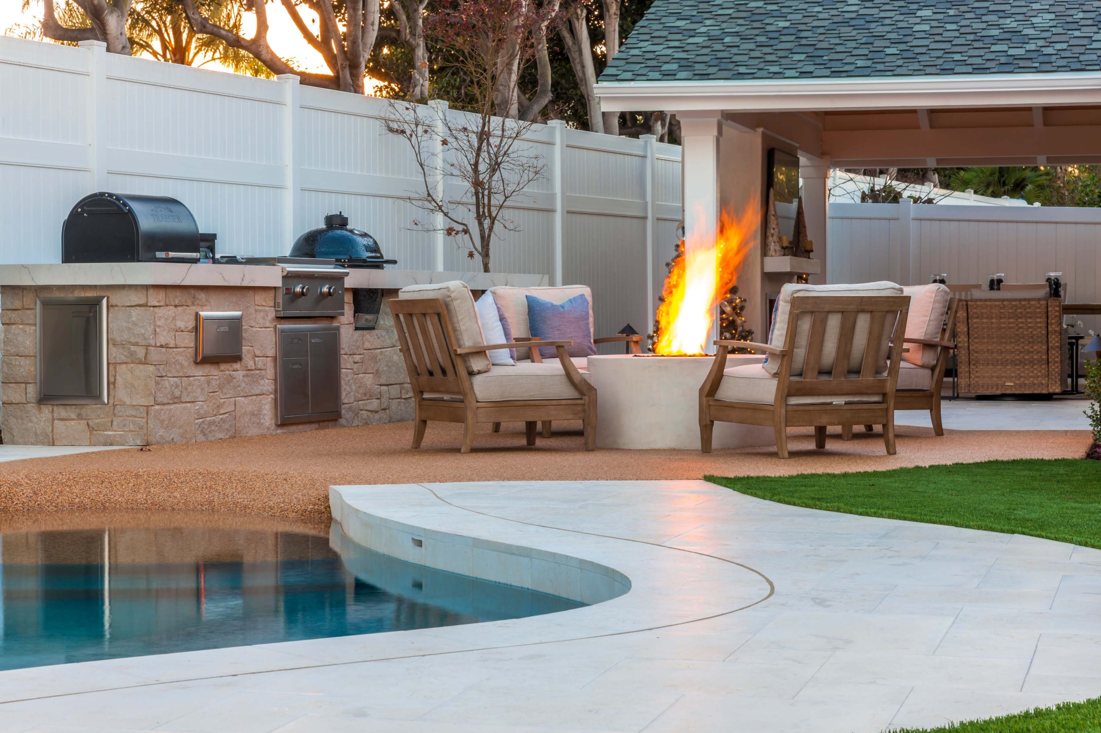 A backyard features a seating area with wooden chairs around a fire pit, an outdoor kitchen, and a pool with a curved edge.