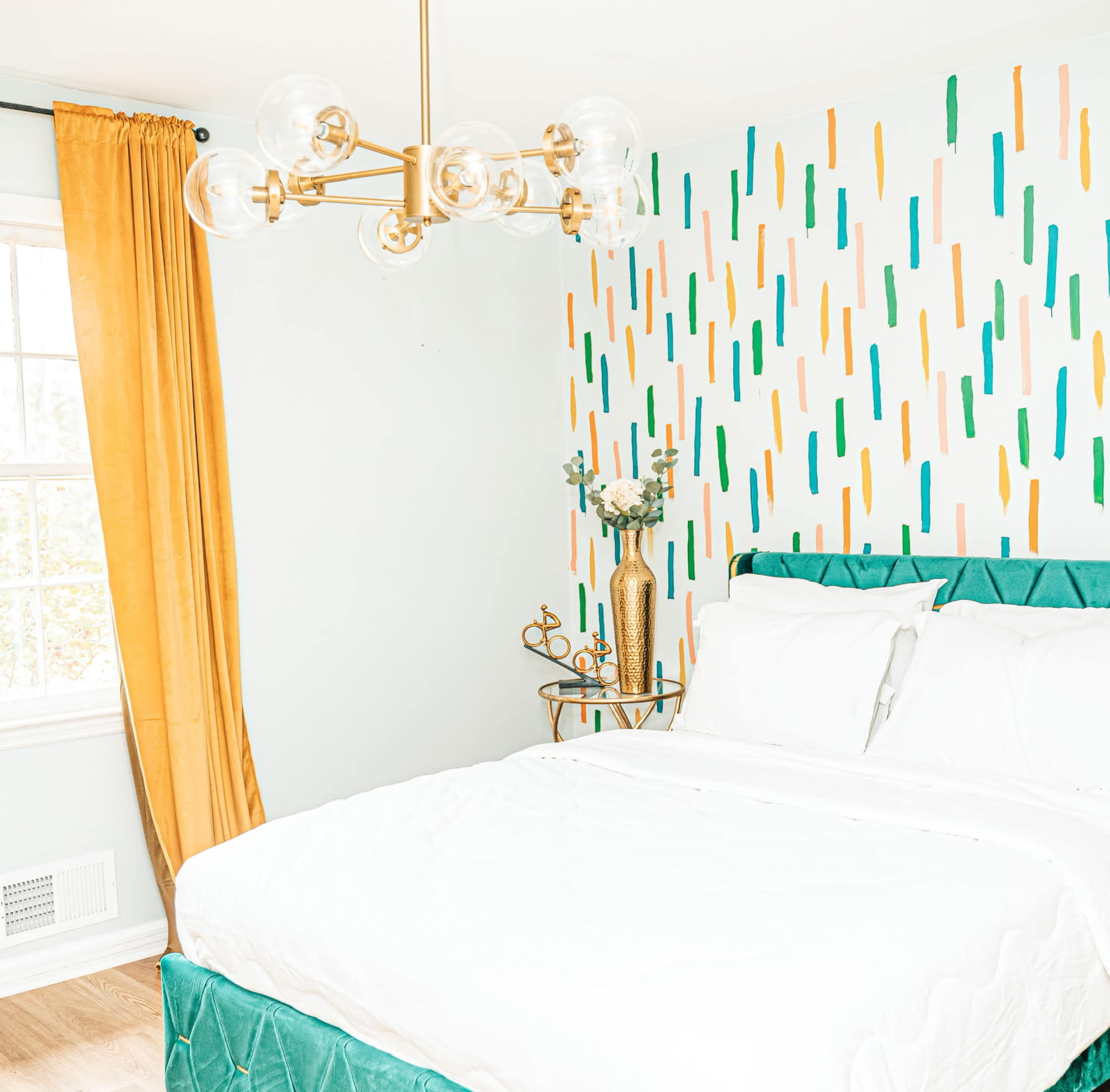 A brightly lit bedroom featuring a bed with white linens, a patterned accent wall, a gold light fixture, and a curtain made of mustard-colored fabric.