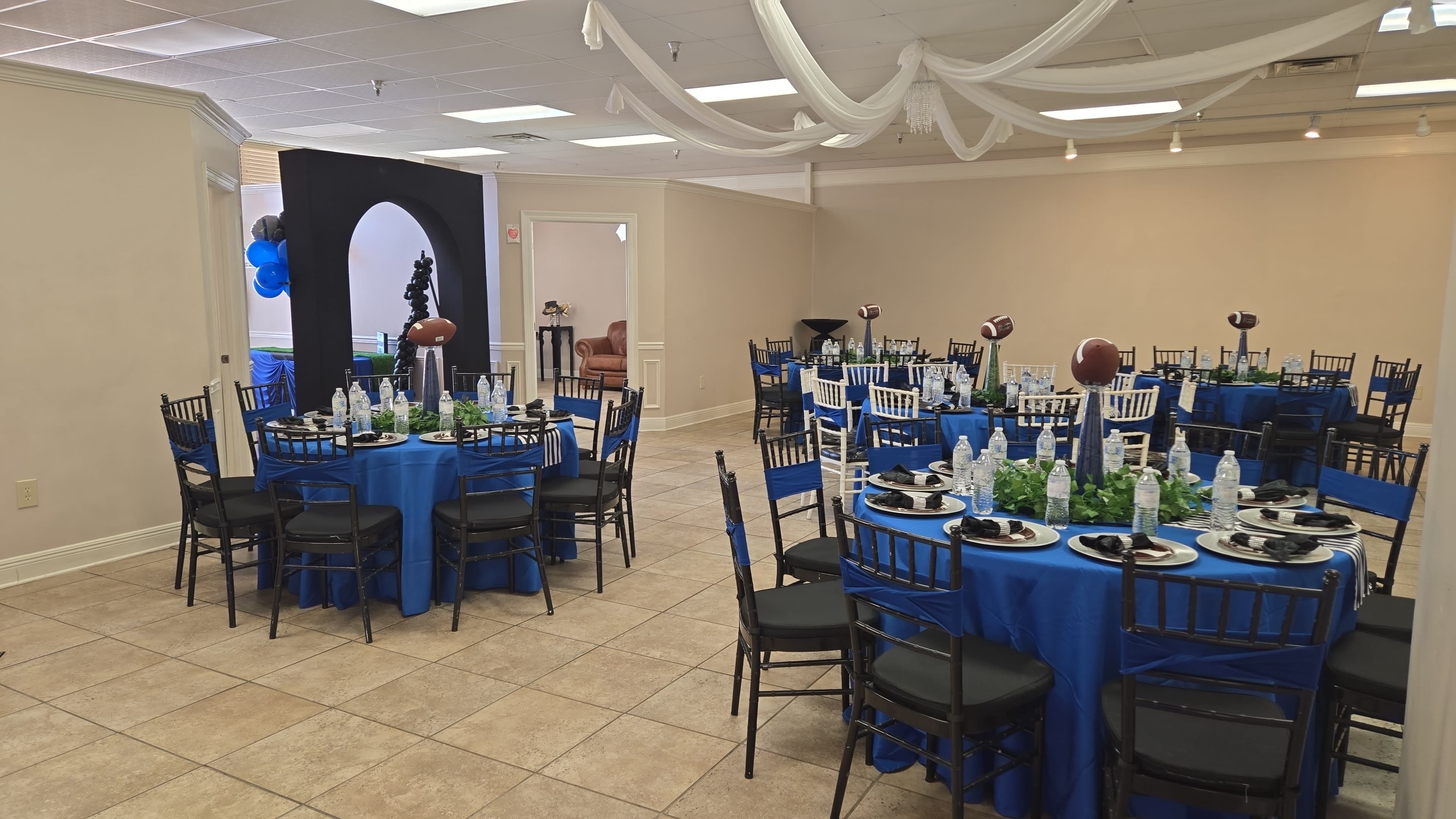 The image shows a party venue with several round tables covered in blue tablecloths, each set with black chairs and decorated with green centerpieces and football-themed accessories.