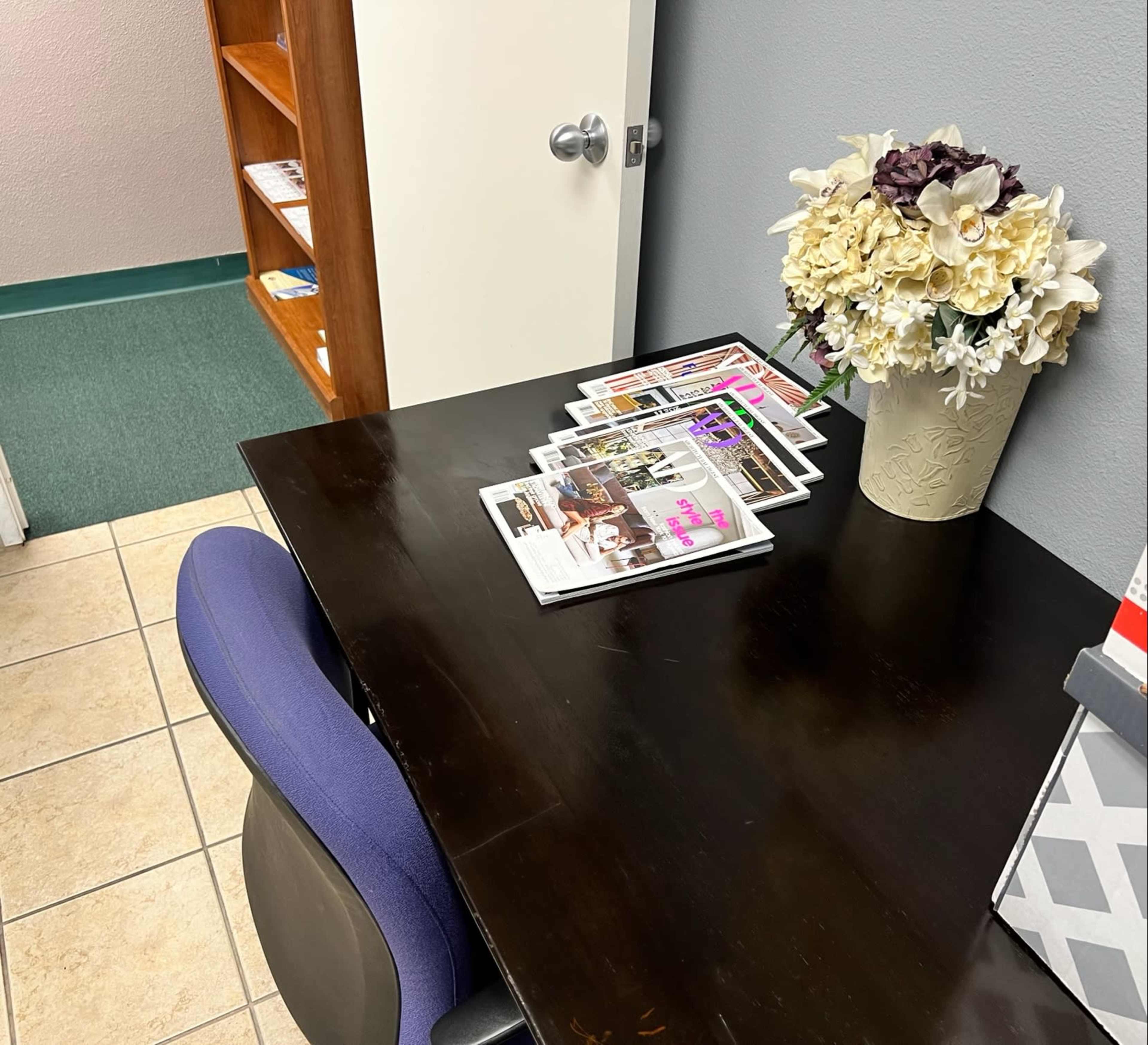 A dark wooden desk with a blue chair holds several magazines, next to a vase of artificial flowers, with a bookshelf visible in the background.