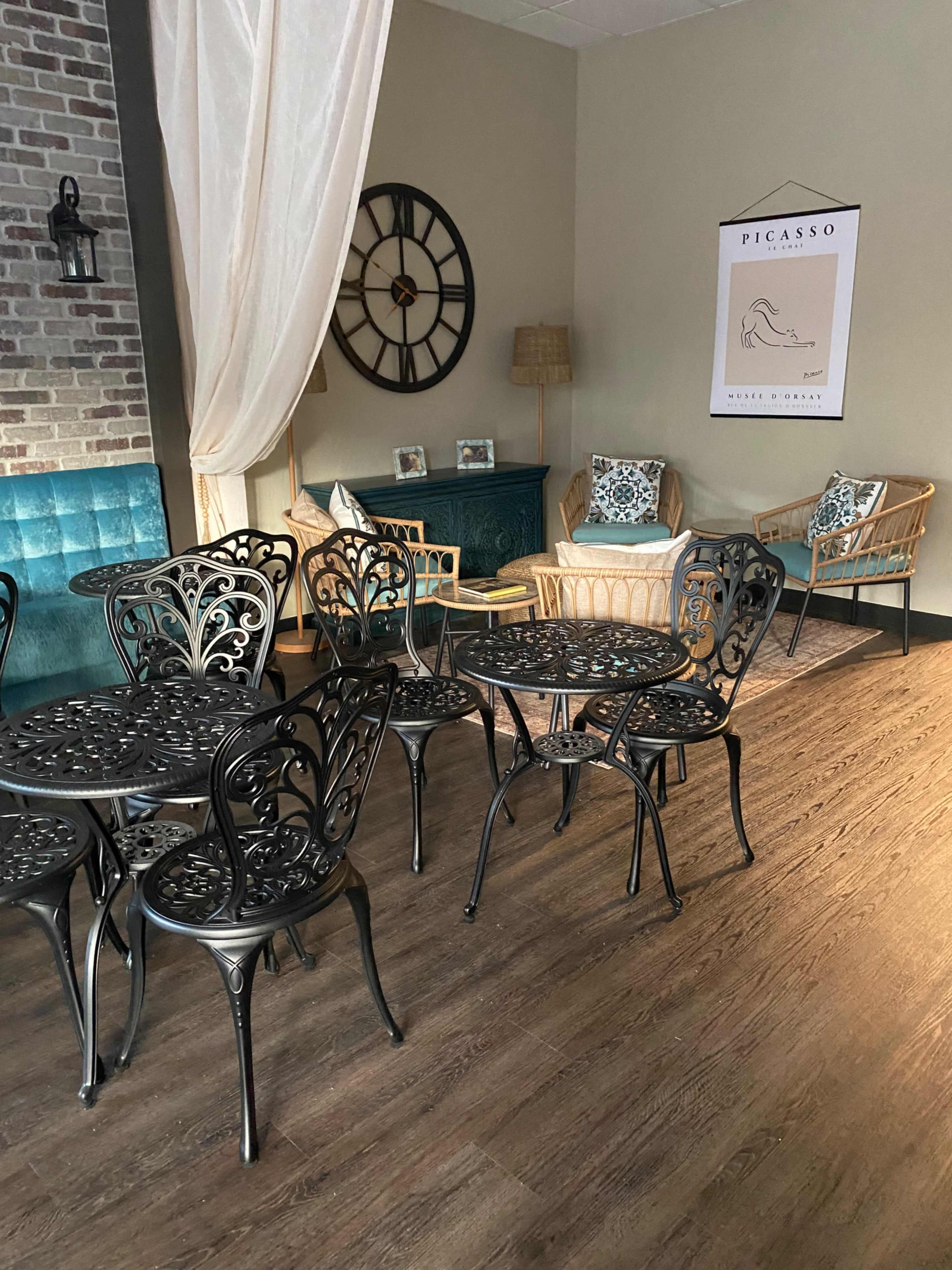 A cozy café interior with wrought iron tables and chairs, a blue cushioned seating area, and a wall adorned with a clock and framed artwork.